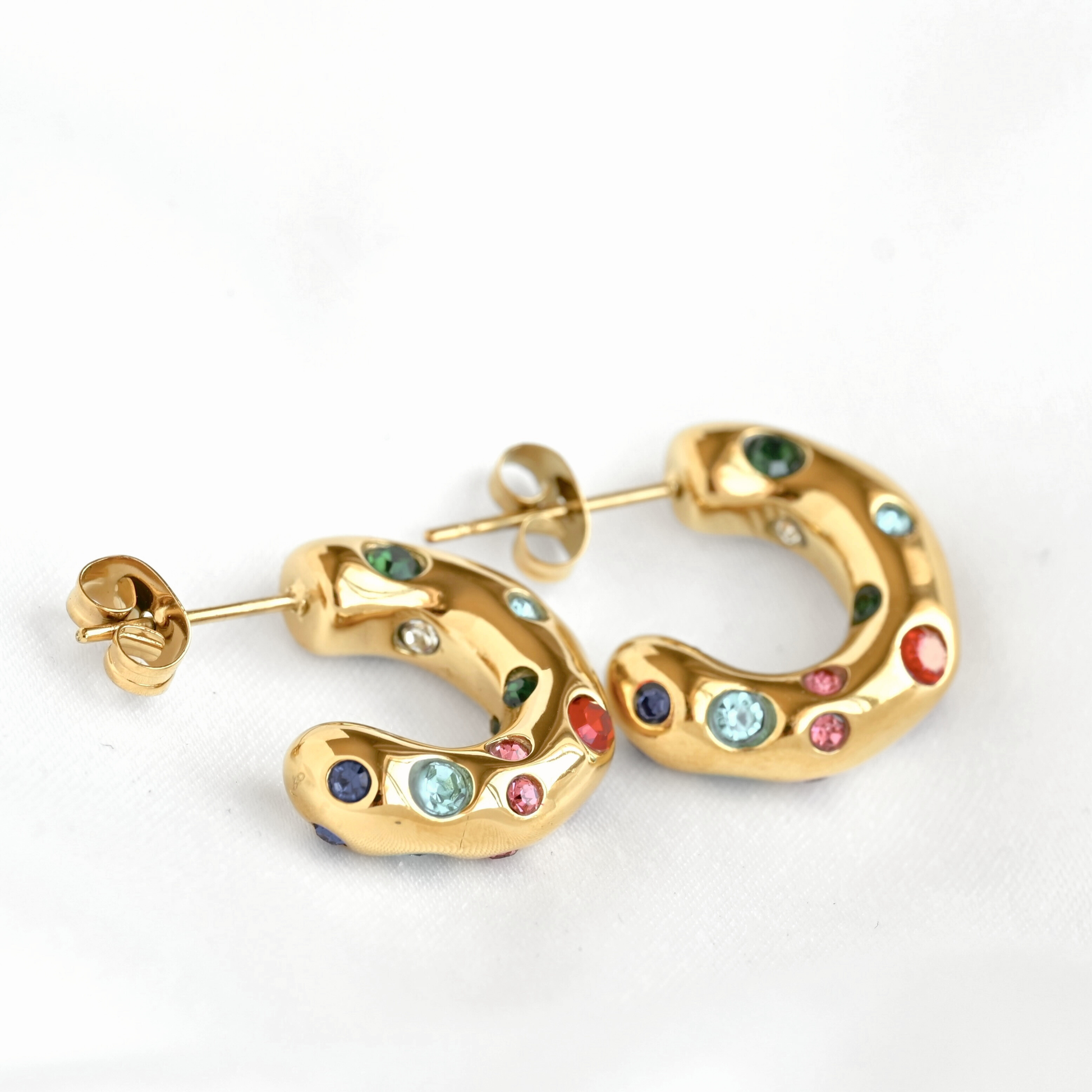 Gold hoop earrings with curved organic shapes, decorated with multiple small colorful gemstones in green, blue, pink, red, and dark blue, shown on a white background.