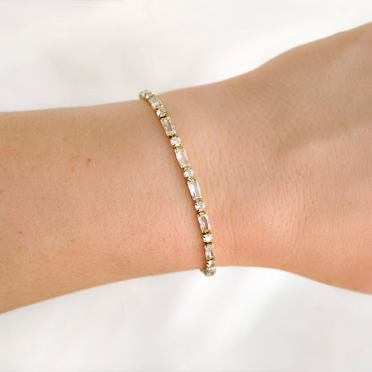 A delicate tennis bracelet with baguette-cut clear crystals set in a yellow gold band, shown worn on a person's wrist against a soft white background.