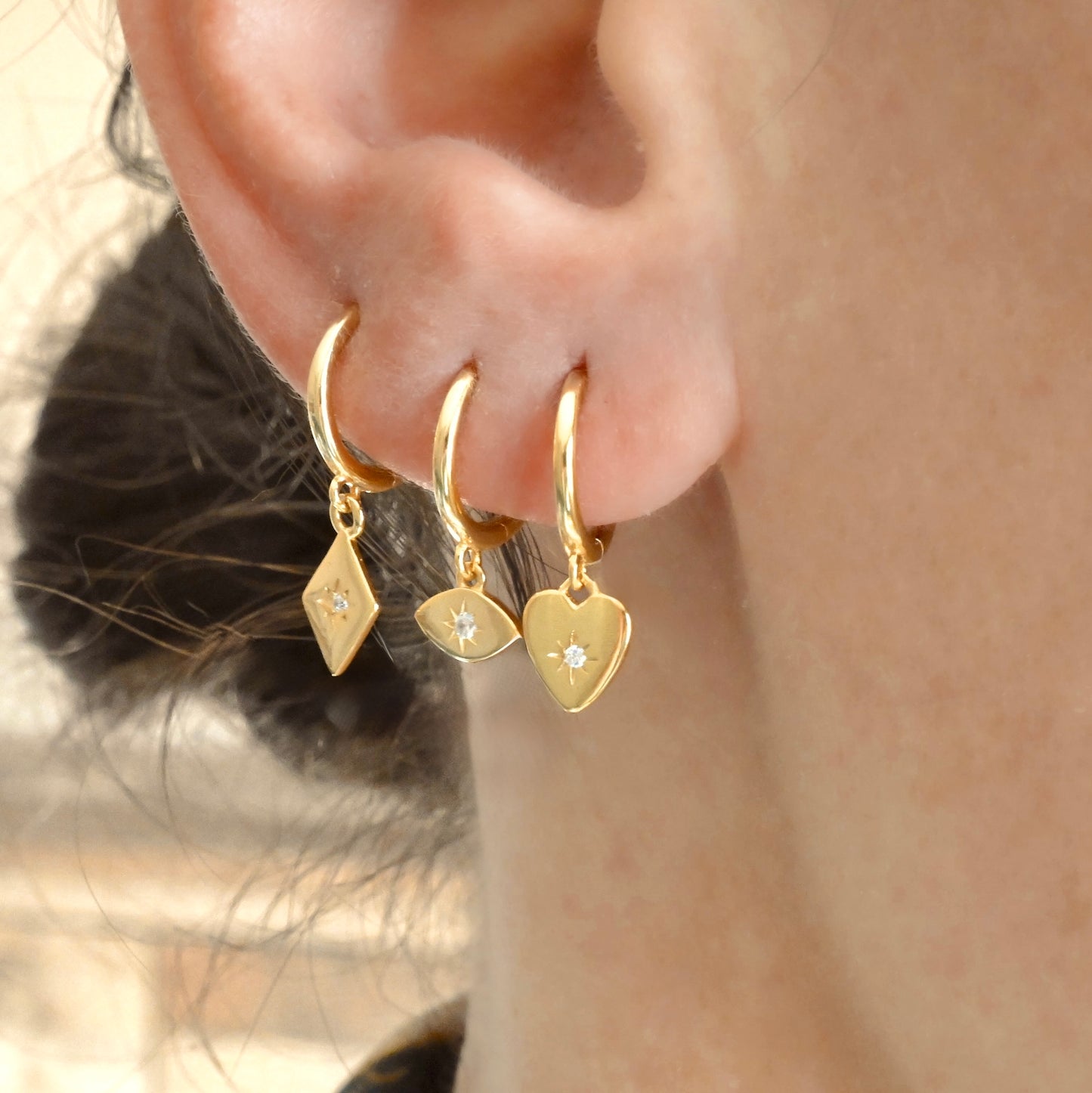 Close-up of a person wearing three gold hoop earrings with heart, evil eye and diamond-shaped charms.