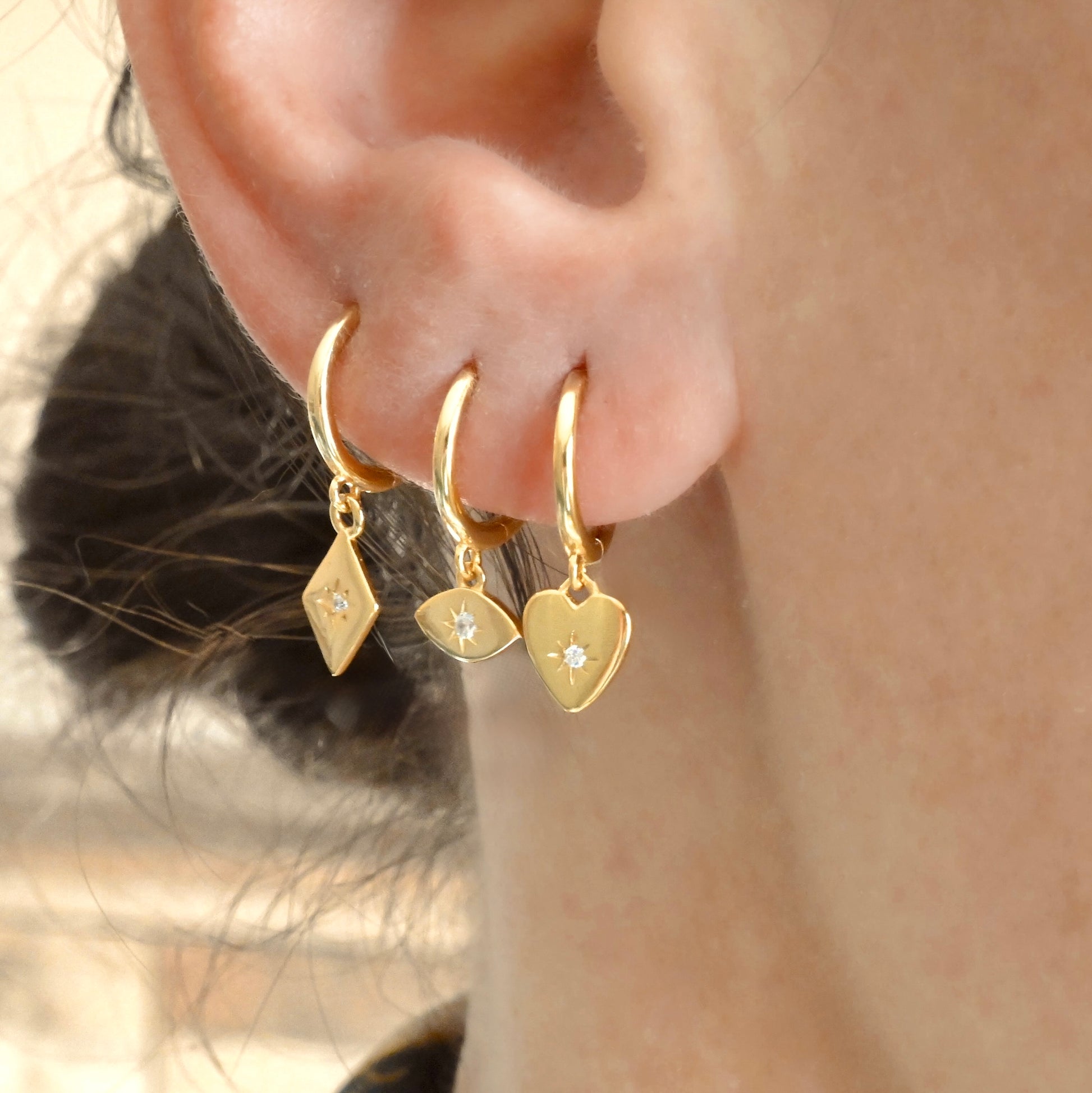 Close-up of a person wearing three gold hoop earrings with heart, evil eye and diamond-shaped charms.