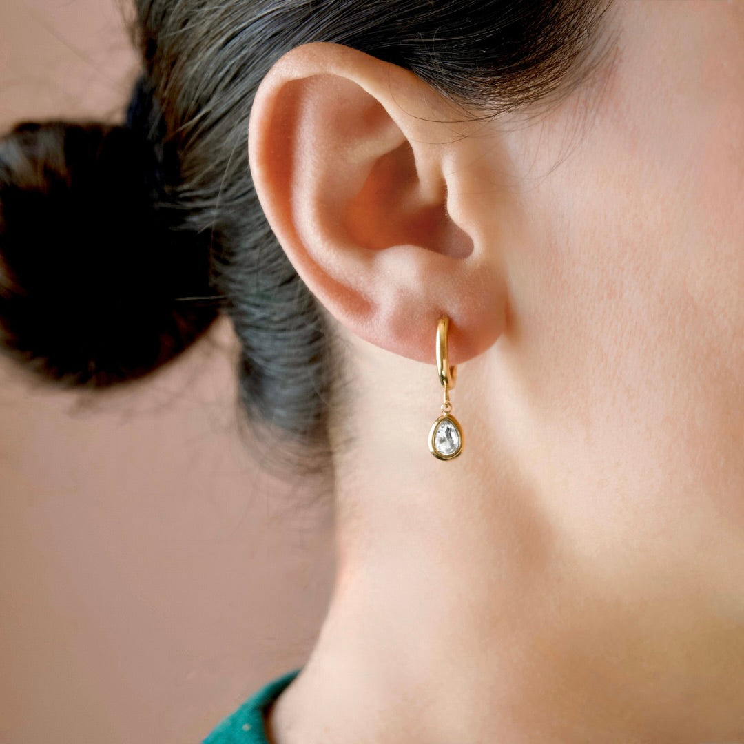 A close-up of a person's ear wearing a small gold huggie hoop earring with a teardrop-shaped clear gemstone drop.