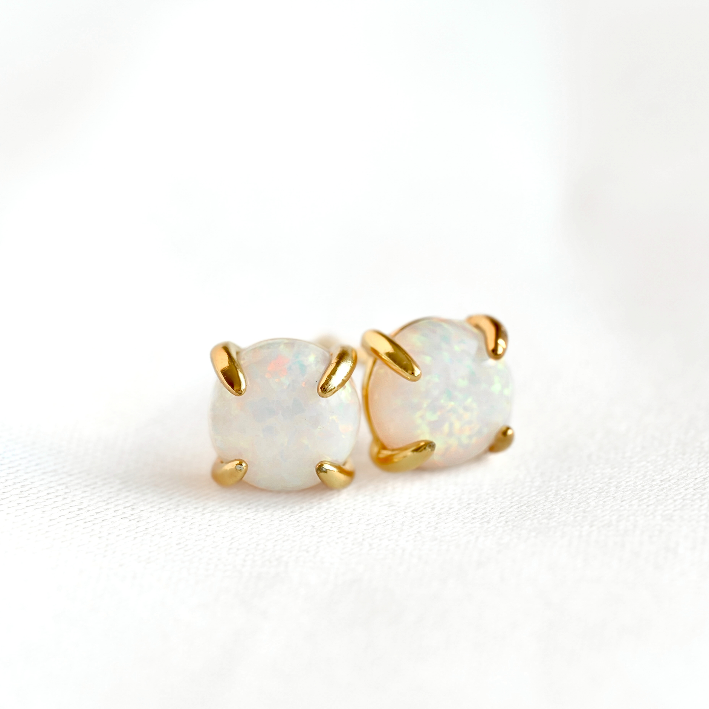 A pair of gold stud earrings with round white opal gemstones, photographed on a clean white textured background.