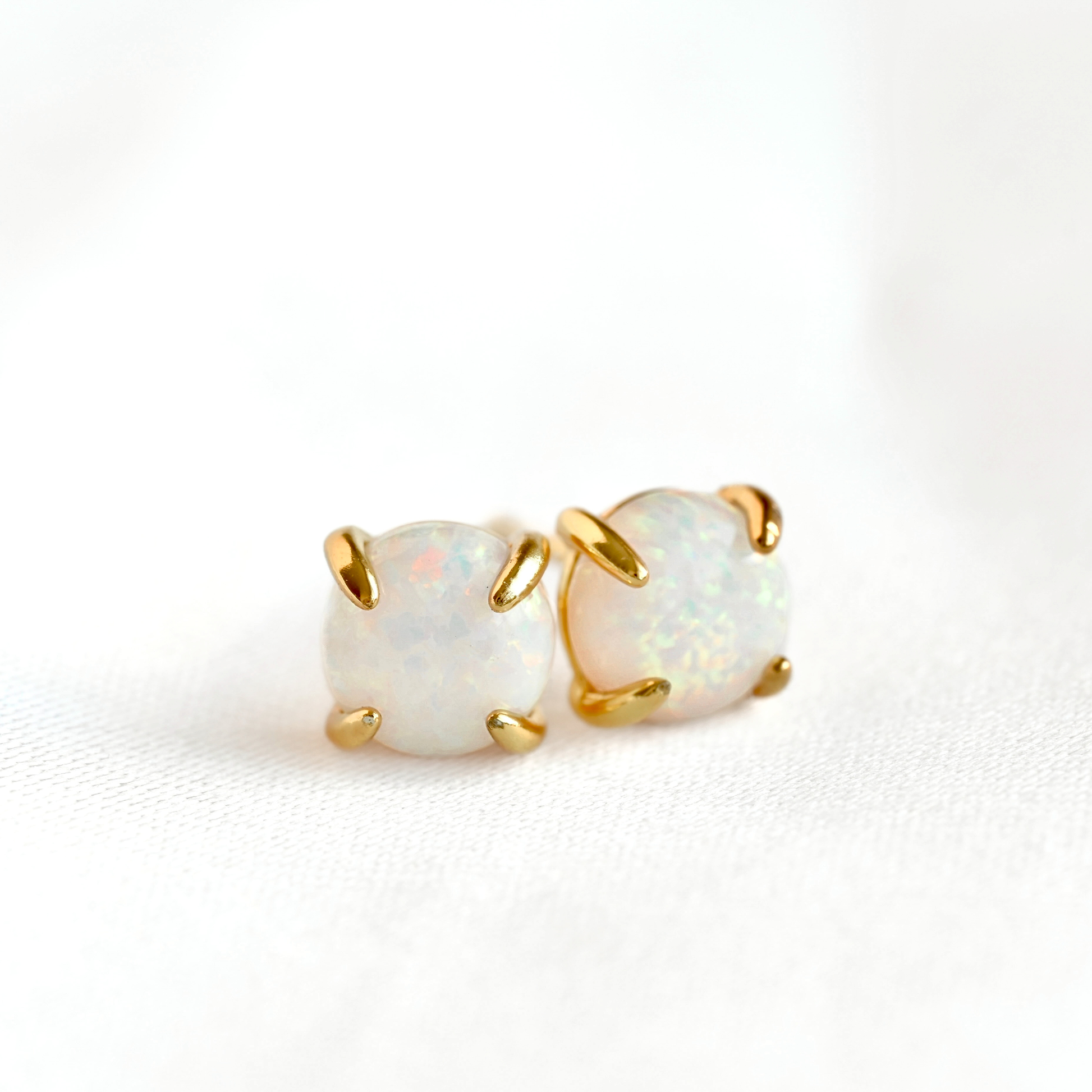 A pair of gold stud earrings with round white opal gemstones, photographed on a clean white textured background.