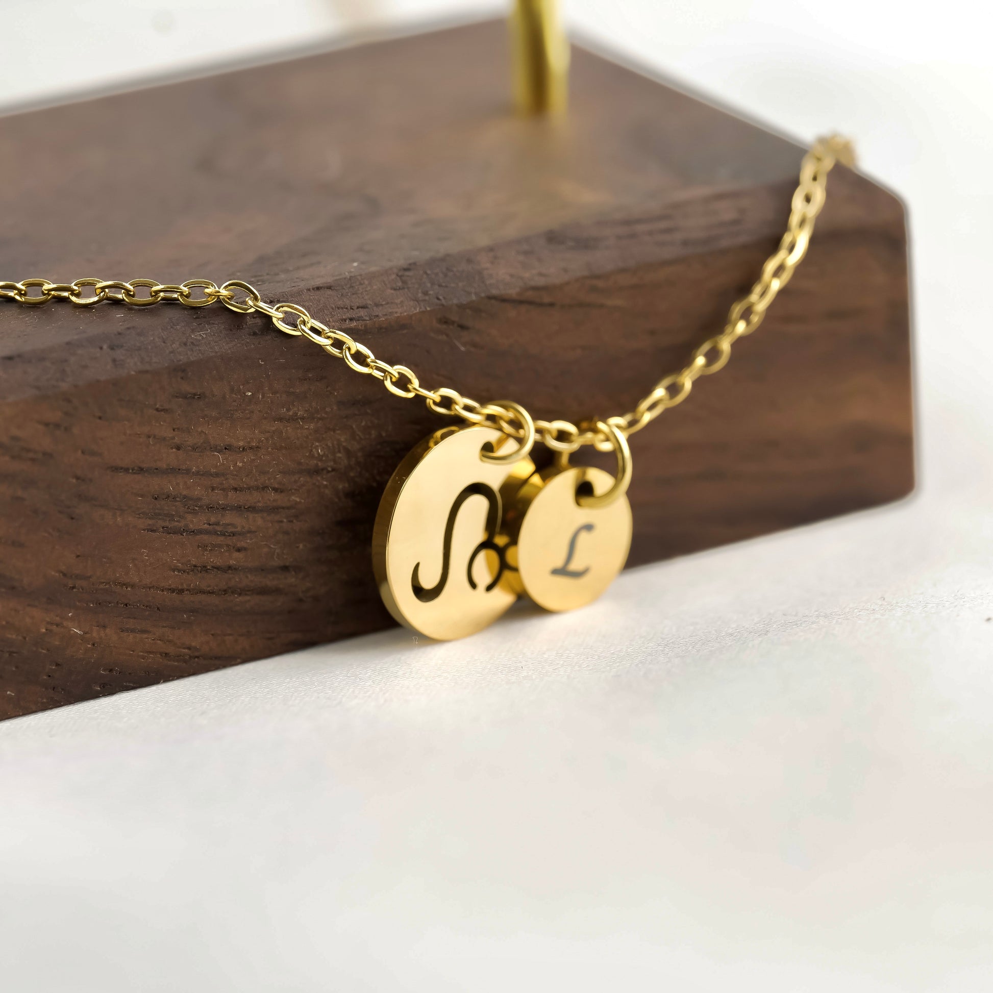 Gold necklace with a coin pendant with 'Leo' symbol and a smaller initial disc with 'L' on a wooden stand.