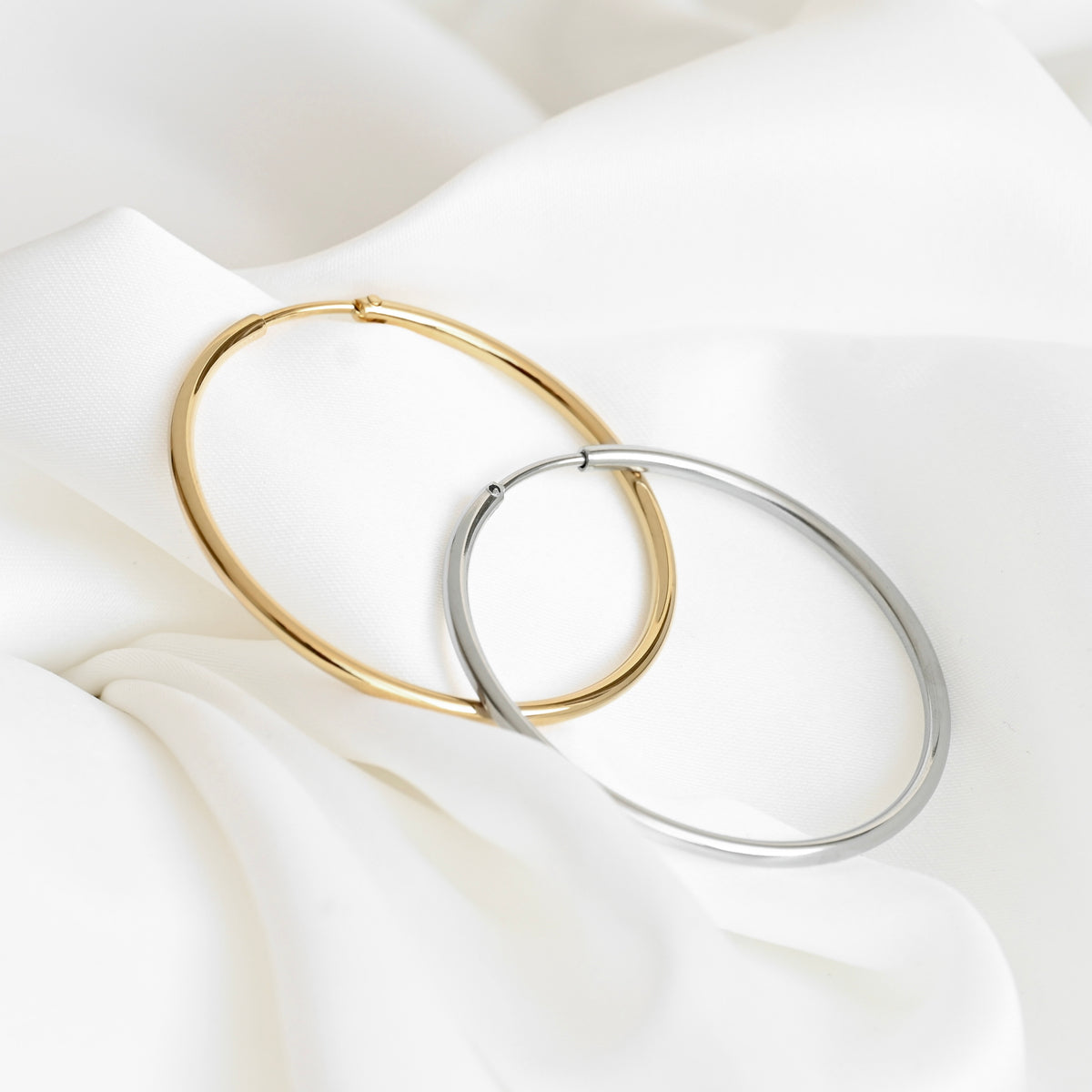 Two large hoop earrings, one in polished gold and one in polished silver, overlapping on a draped white fabric background.