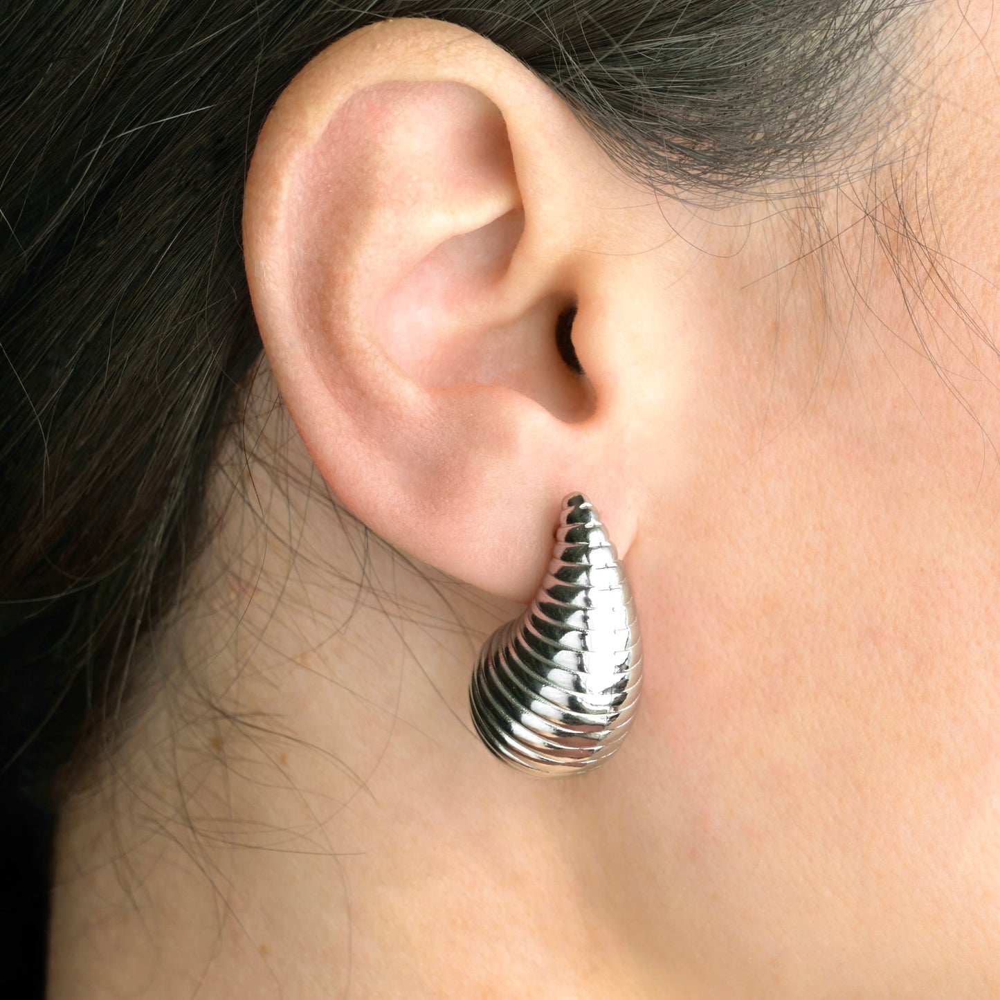 Silver ribbed bottega earrings worn on an ear with a dark background
