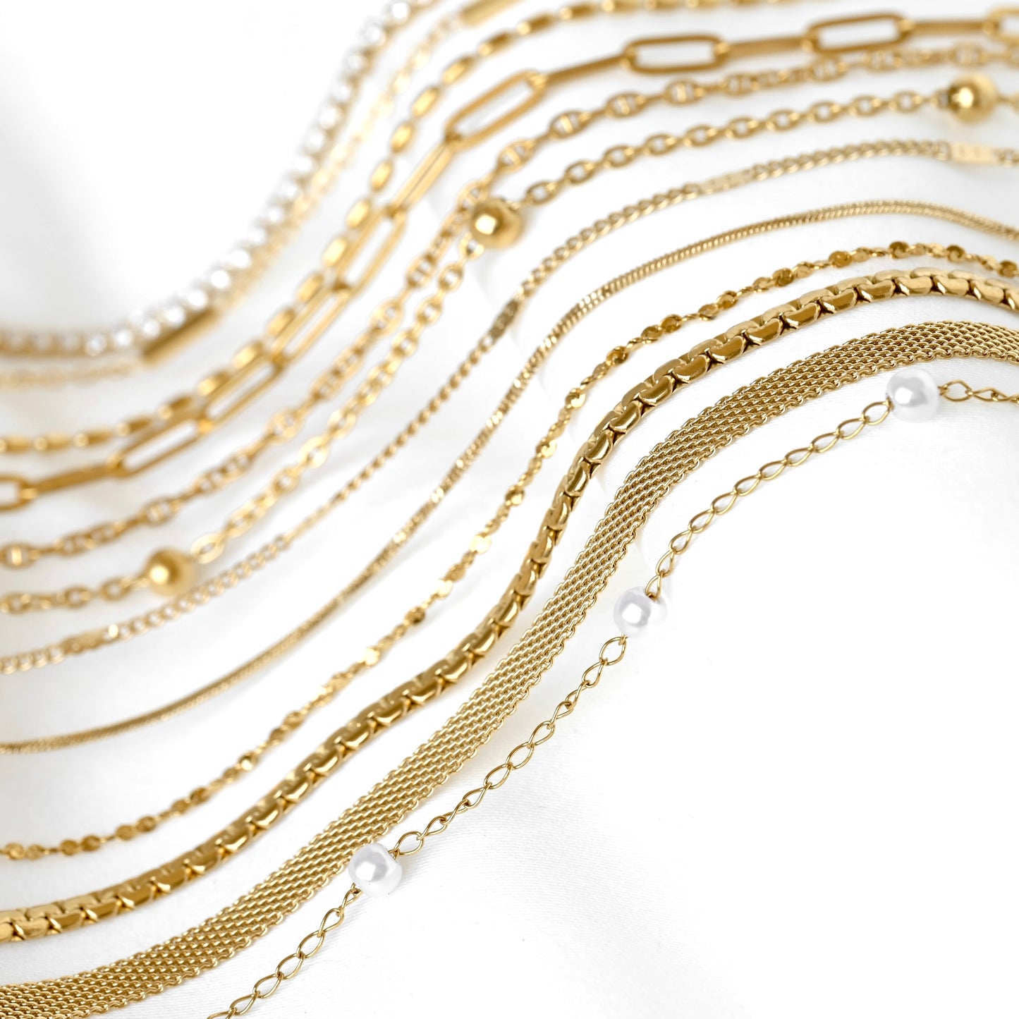 A close-up arrangement of multiple gold-tone and pearl-bead chains draped in curved rows on a white background.
