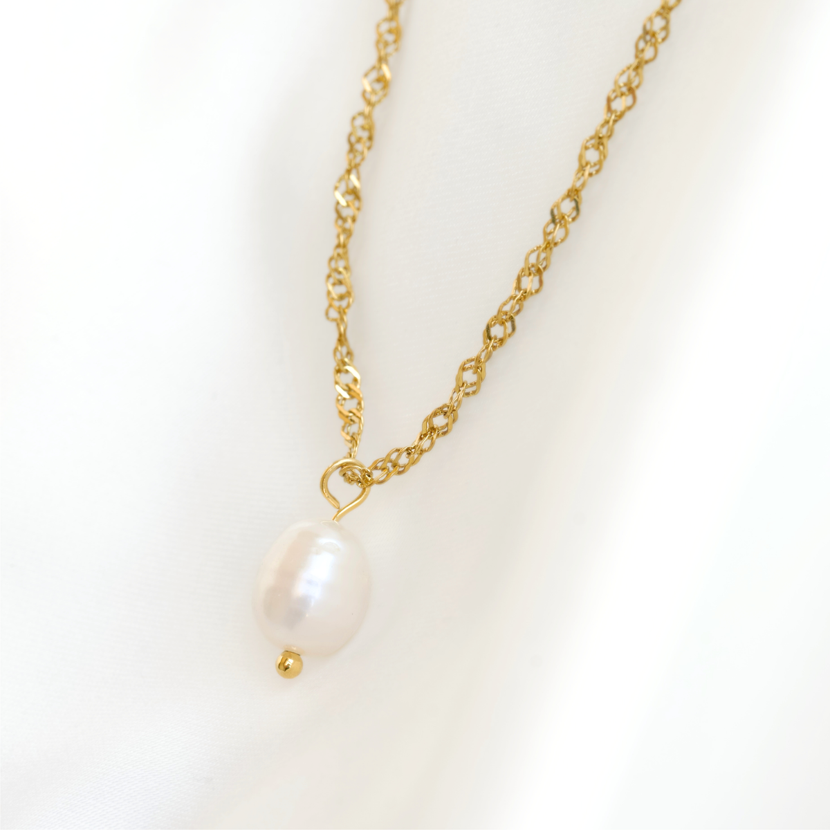 A delicate gold chain necklace featuring a single white baroque pearl pendant, displayed on a soft white fabric background.