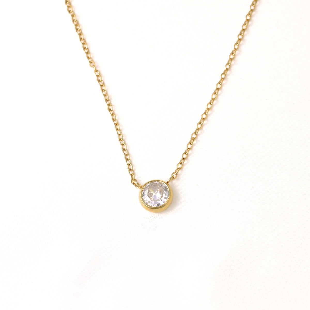 A delicate gold chain necklace featuring a small round bezel-set clear gemstone pendant, displayed against a clean white background.
