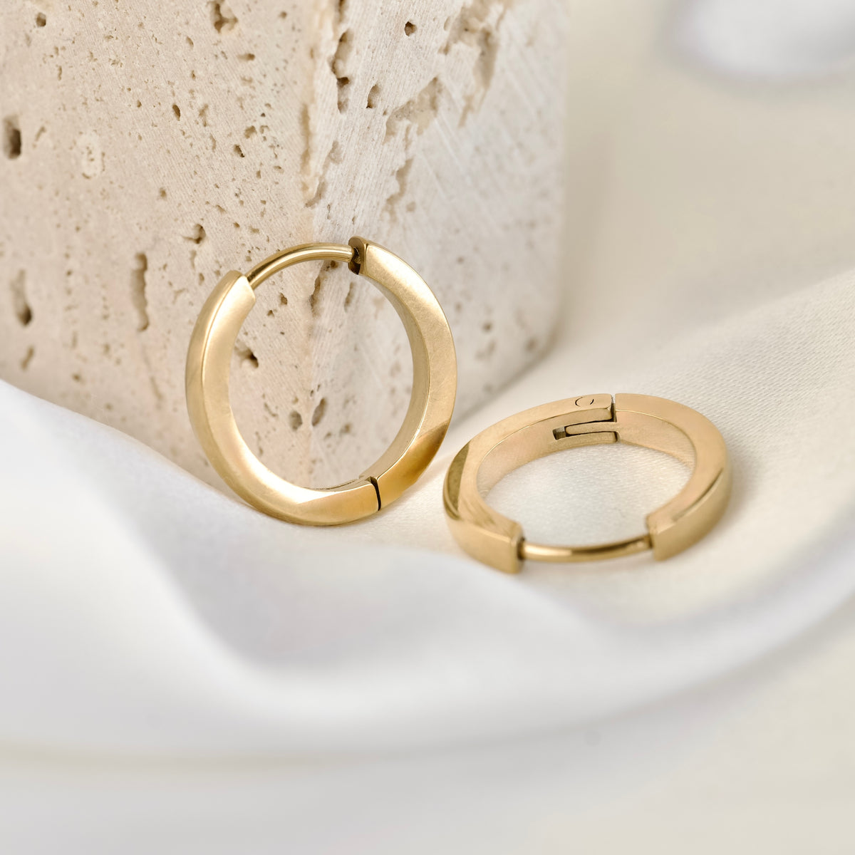 A pair of small polished gold hoop earrings, one resting on white satin and other resting against beige stone.