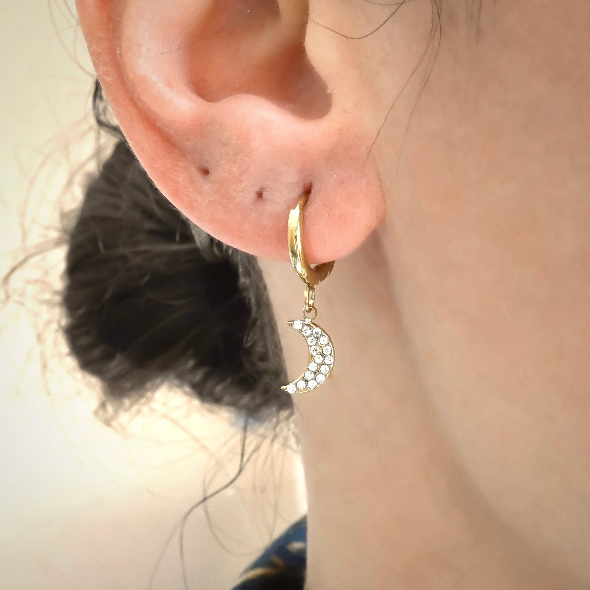 Close-up view of gold huggie hoops with a moon charm with embedded white stones on a person's ear