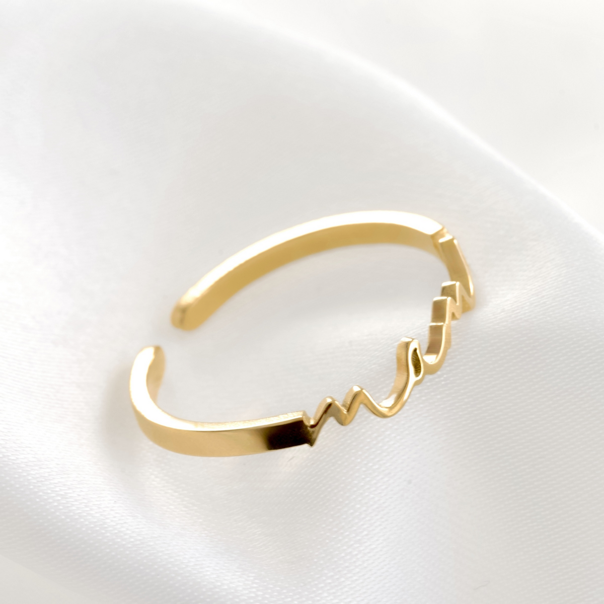 A gold-plated open cuff ring featuring a wavy, calligraphic line detail with 'mama' script, displayed on a soft white fabric background.