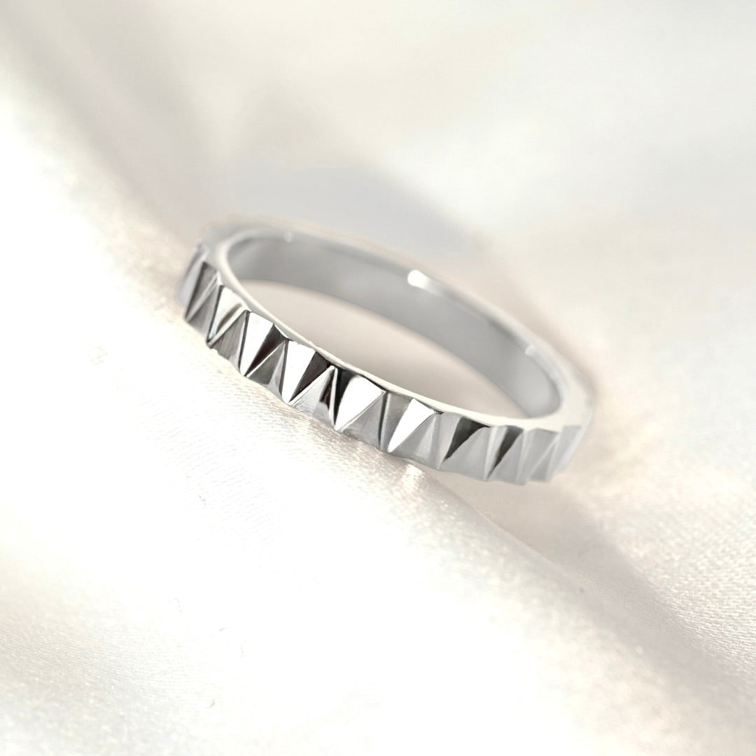A silver pyramid-patterned band ring resting on a soft, white textured fabric background.