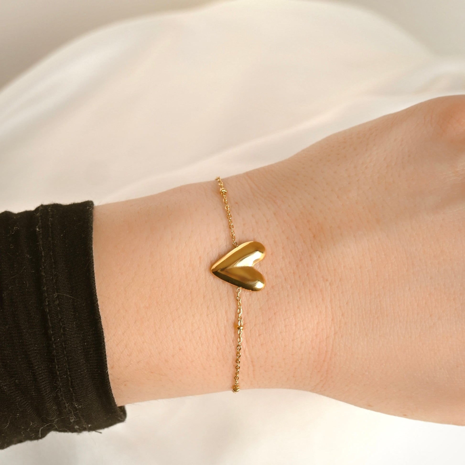 A close-up of a delicate gold chain bracelet with a polished, three-dimensional heart charm worn on a person's wrist.