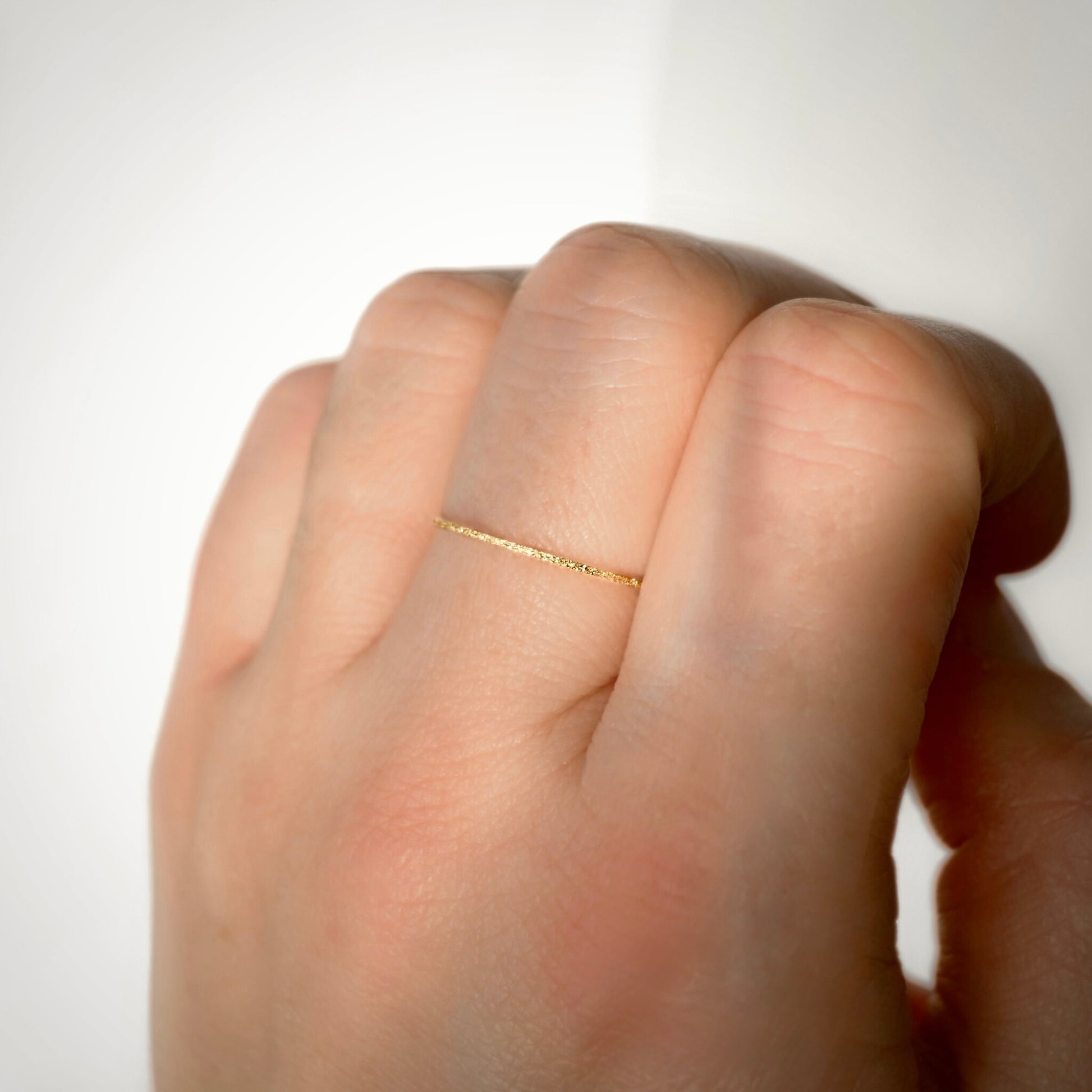 A close-up of a hand wearing a very thin, textured gold band ring against a plain white background.