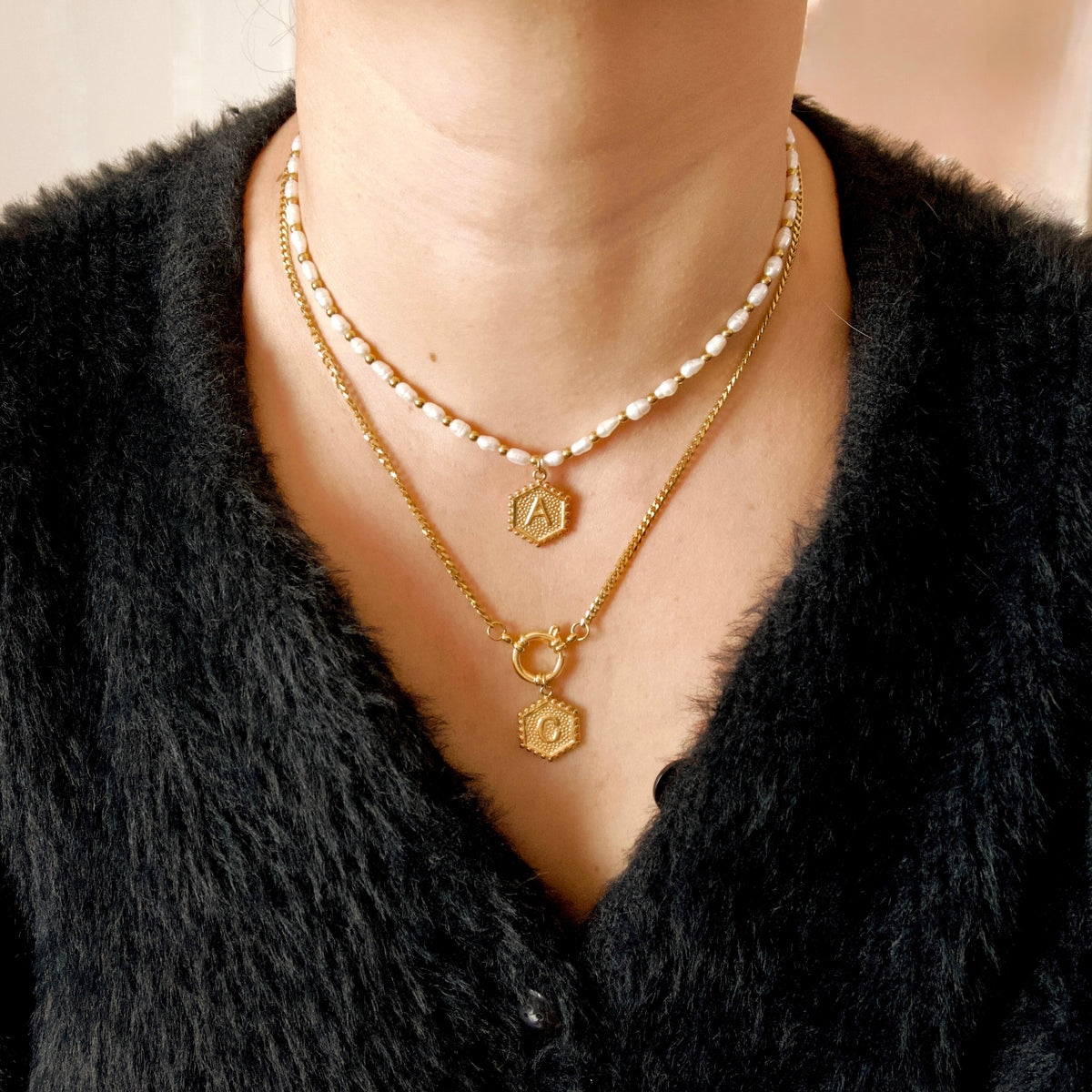 A close-up of a person wearing layered necklaces: a short strand of white pearls, a gold chain with a small hexagonal pendant engraved with the letter A, and a longer gold chain with a ring connector and hexagonal pendant engraved with the letter O. The person is wearing a black fuzzy V-neck sweater.