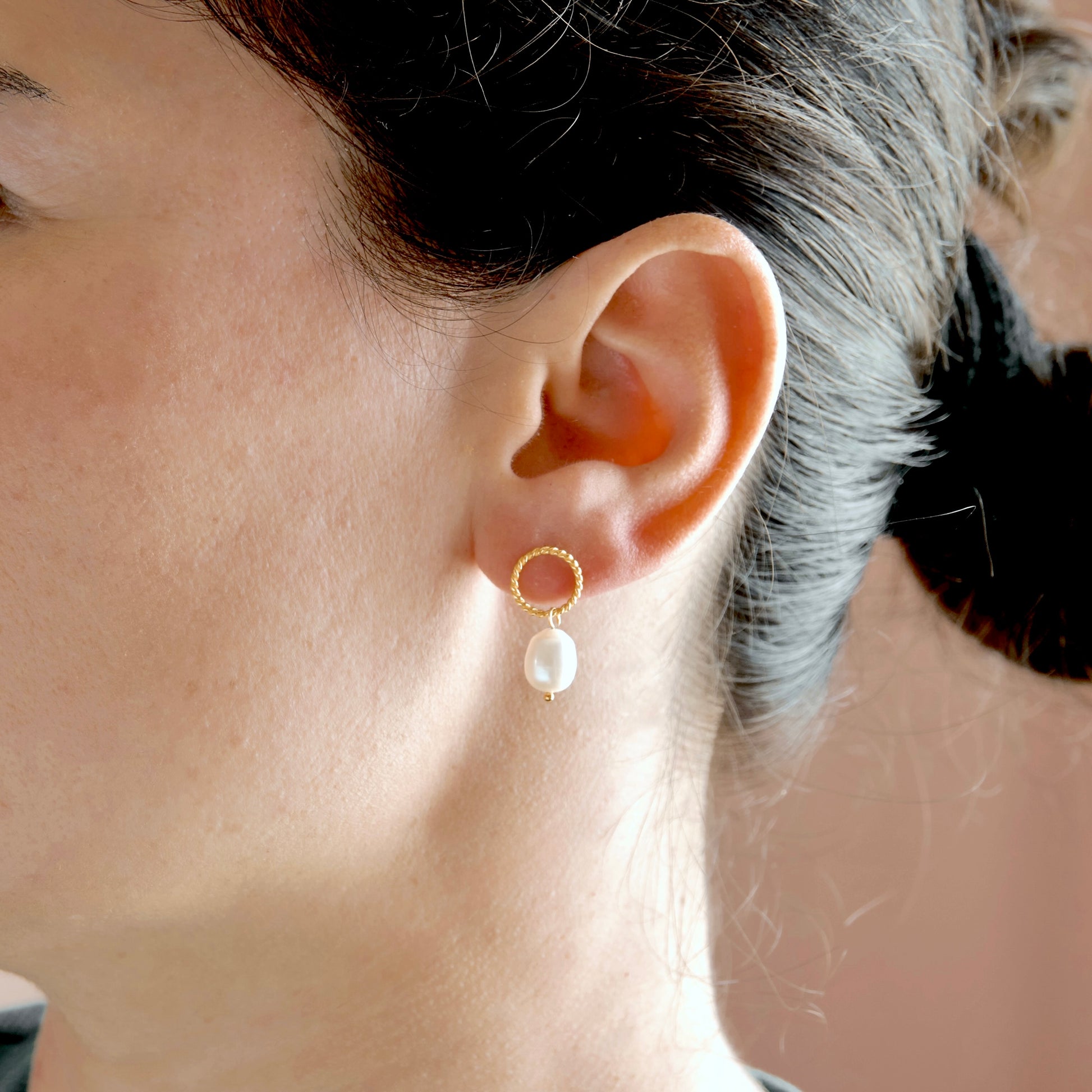 A close-up of a model's ear wearing a delicate gold hoop earring with a dangling white pearl.