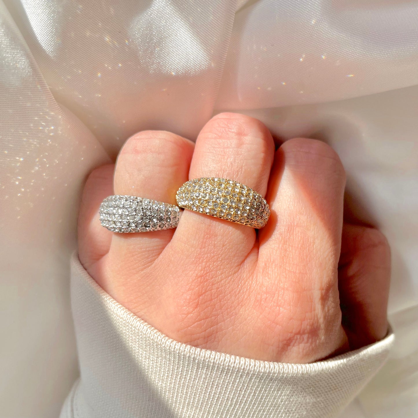 Hand wearing two sparkling diamond rings, one gold and one silver, on a light fabric background under direct sunlight