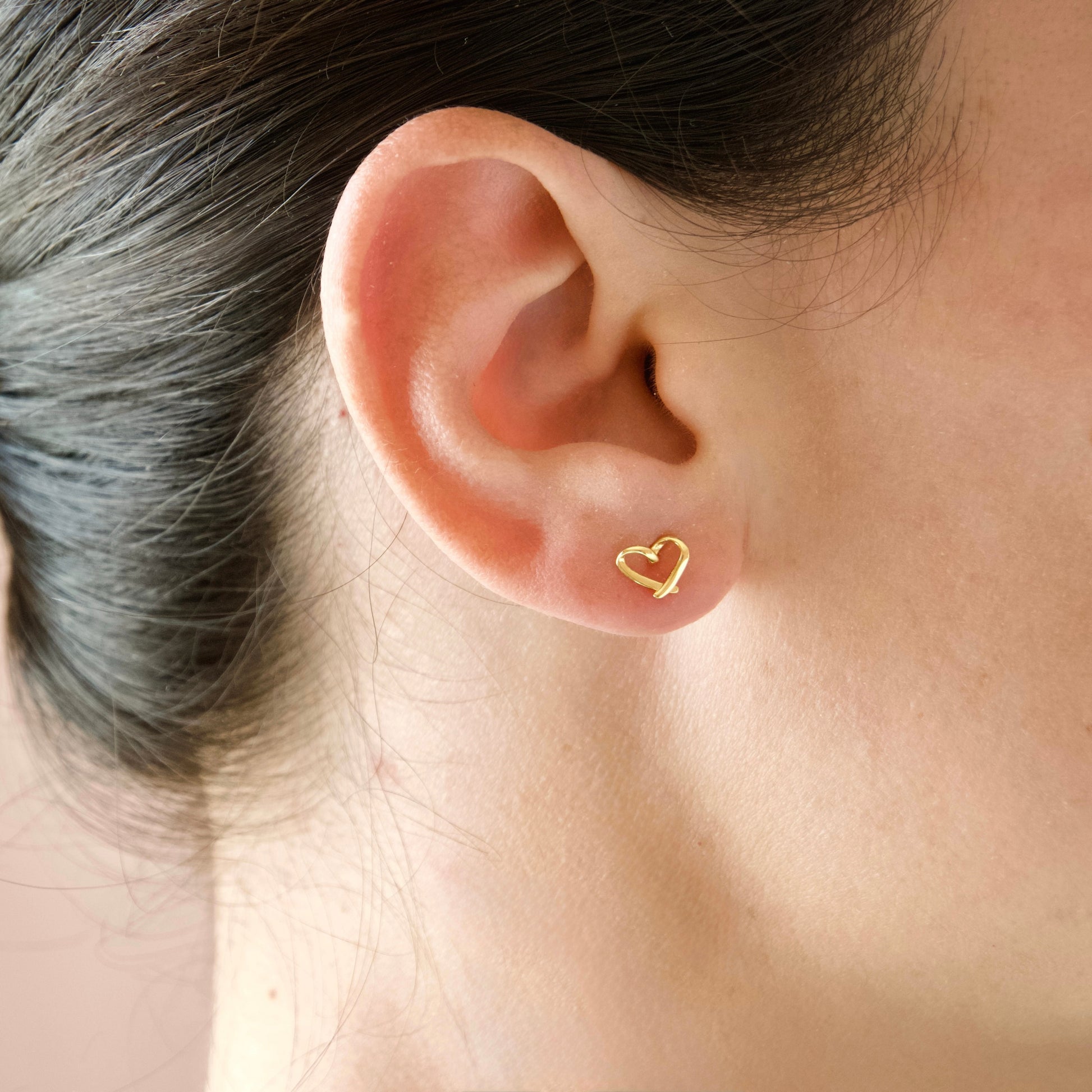 A close-up of a person's ear wearing a small, delicate gold heart-shaped stud earring.