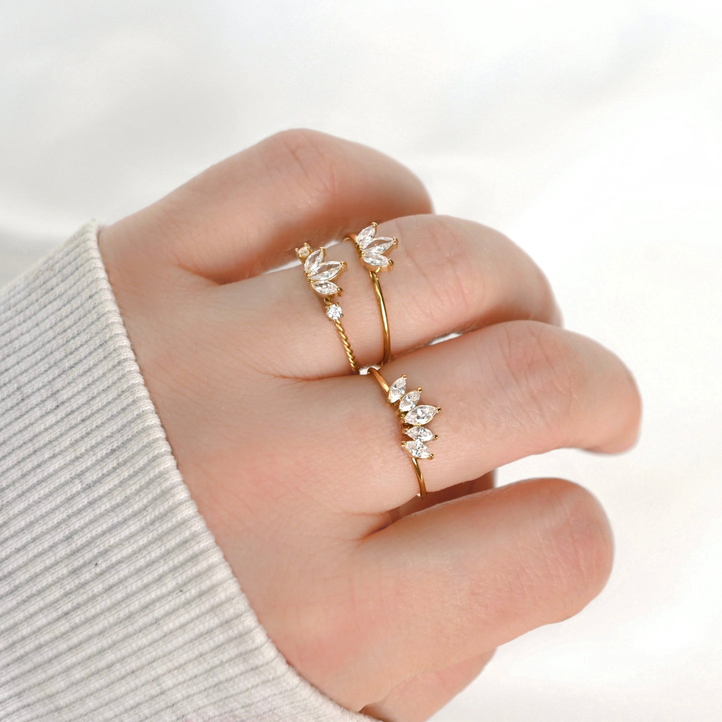 A close-up of a hand wearing three delicate gold rings, each decorated with small clear crystals in different floral and leaf motifs, set against a soft white background.