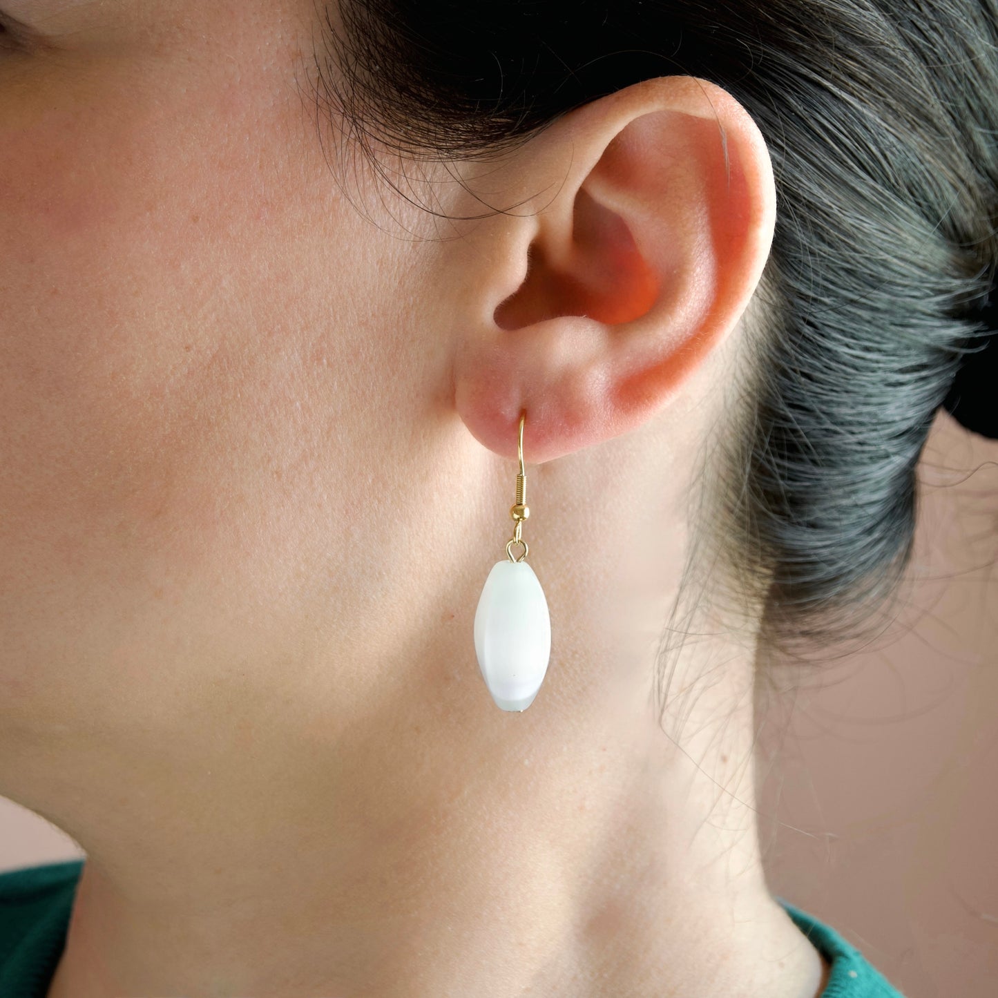 A close-up of a person's ear wearing a single drop earring with a gold hook and a smooth, oval white stone pendant.