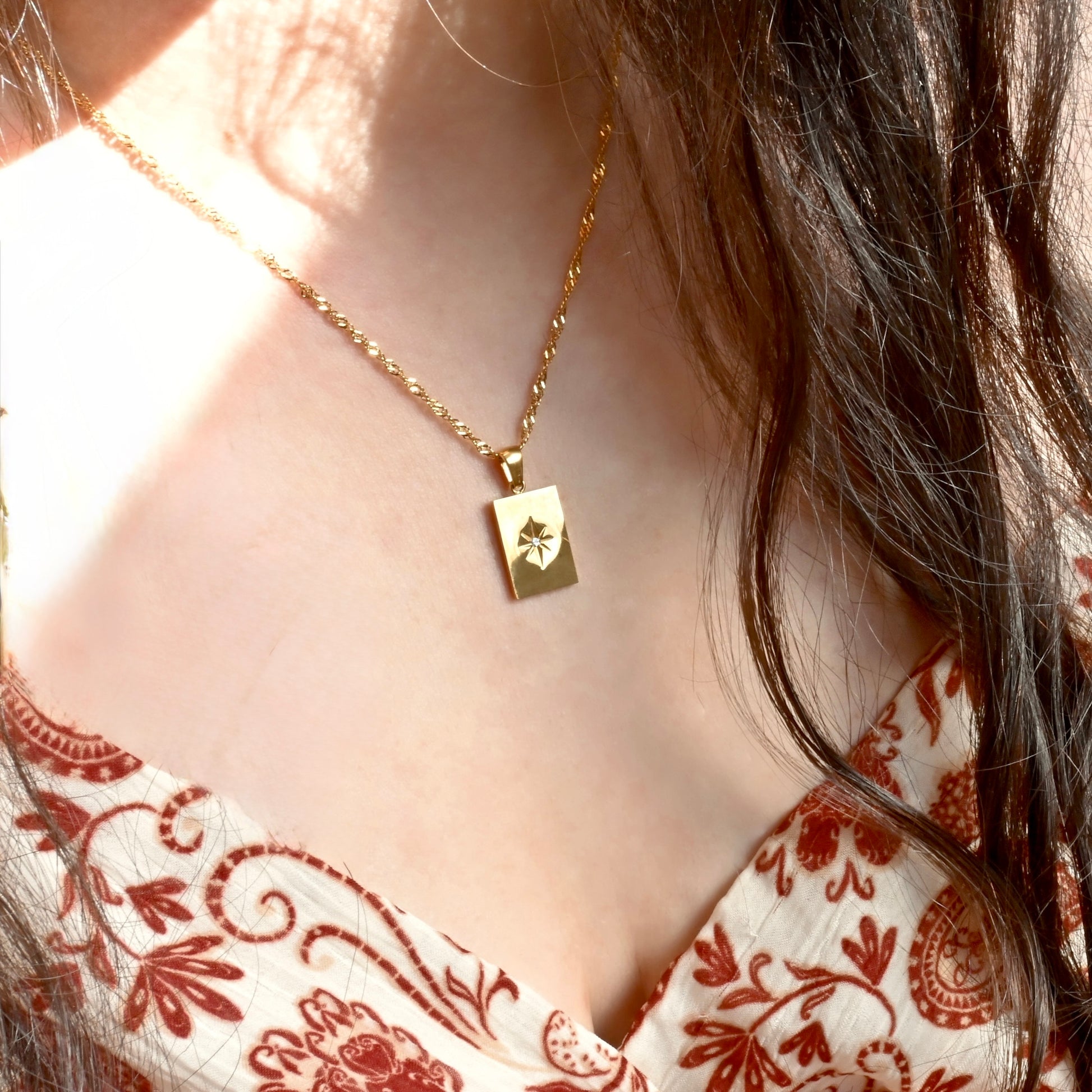 Gold rectangular north star necklace with a twisted chain worn by a person in a patterned top.