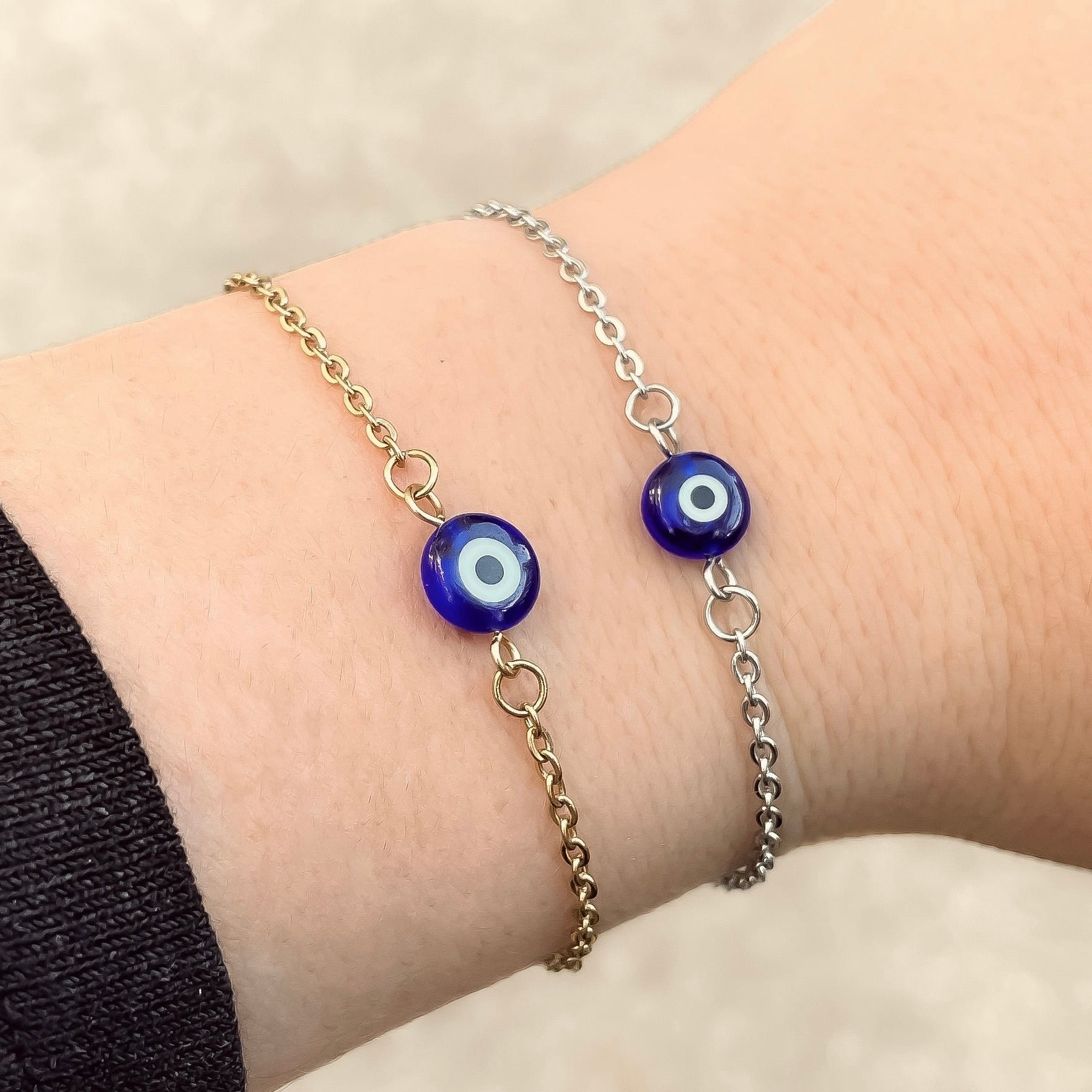 Two bracelets, one gold one silver, with blue evil eye beads on a wrist against a neutral background