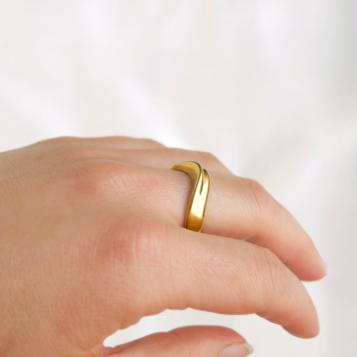 A close-up of a hand wearing a polished, curved gold ring against a soft, light background.