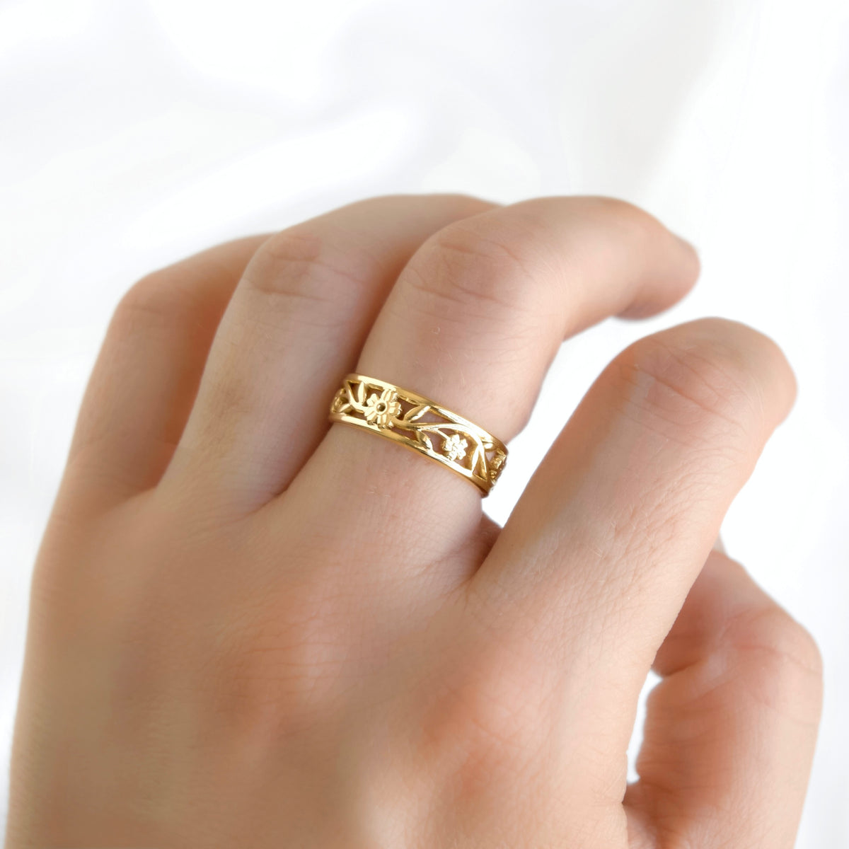 A dainty gold band with cut-out filigree pattern with flowers and leafs on a women's middle finger.