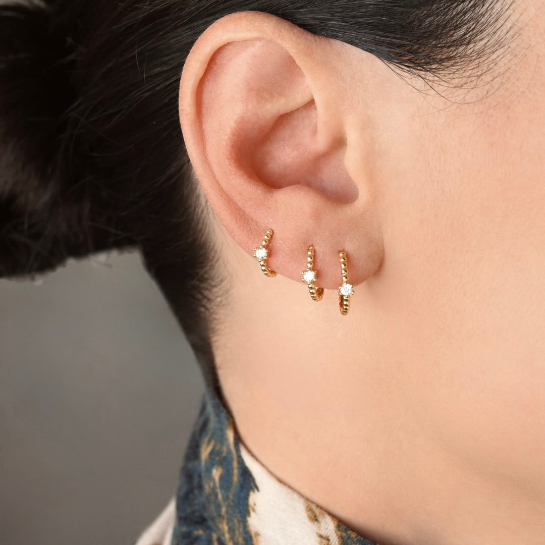 Close-up of a person's ear wearing three small gold beaded huggie hoop earrings, each with a tiny clear stone, against a soft gray background.
