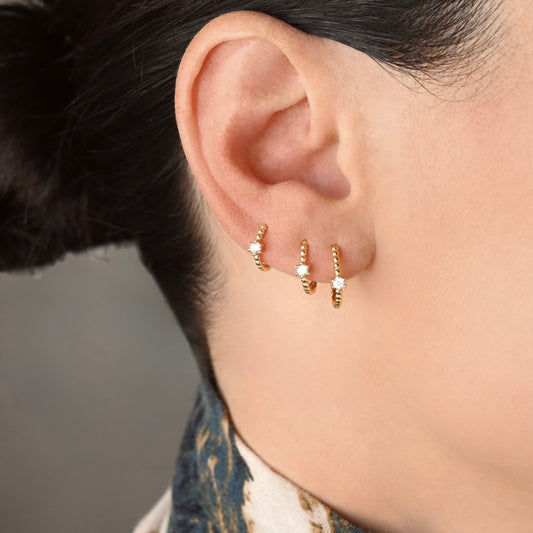 Close-up of a person's ear wearing three small gold beaded huggie hoop earrings, each with a tiny clear stone, against a soft gray background.