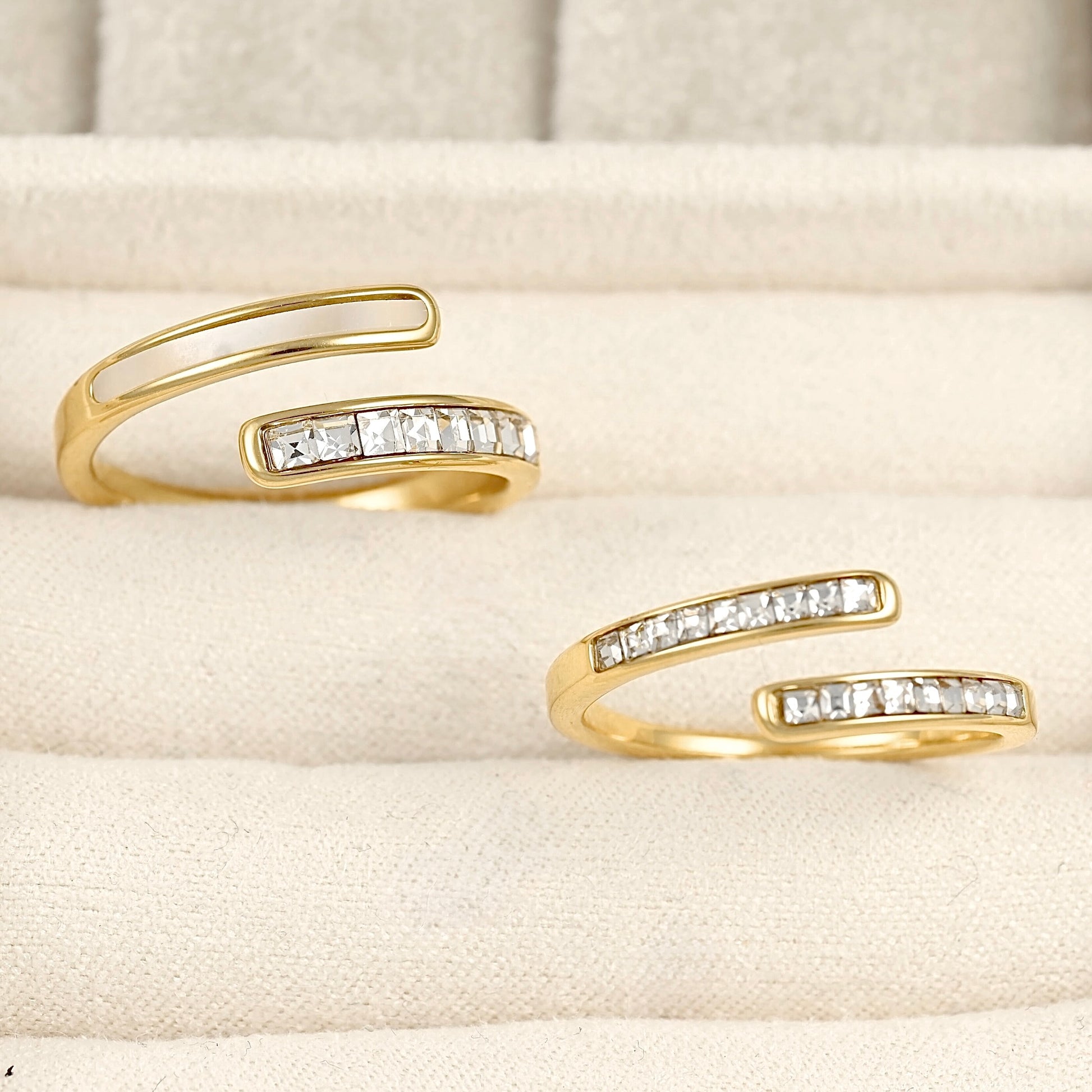 Two gold open wrap rings with a row of clear rectangular CZ stones, displayed on a cream-colored textured jewelry stand.