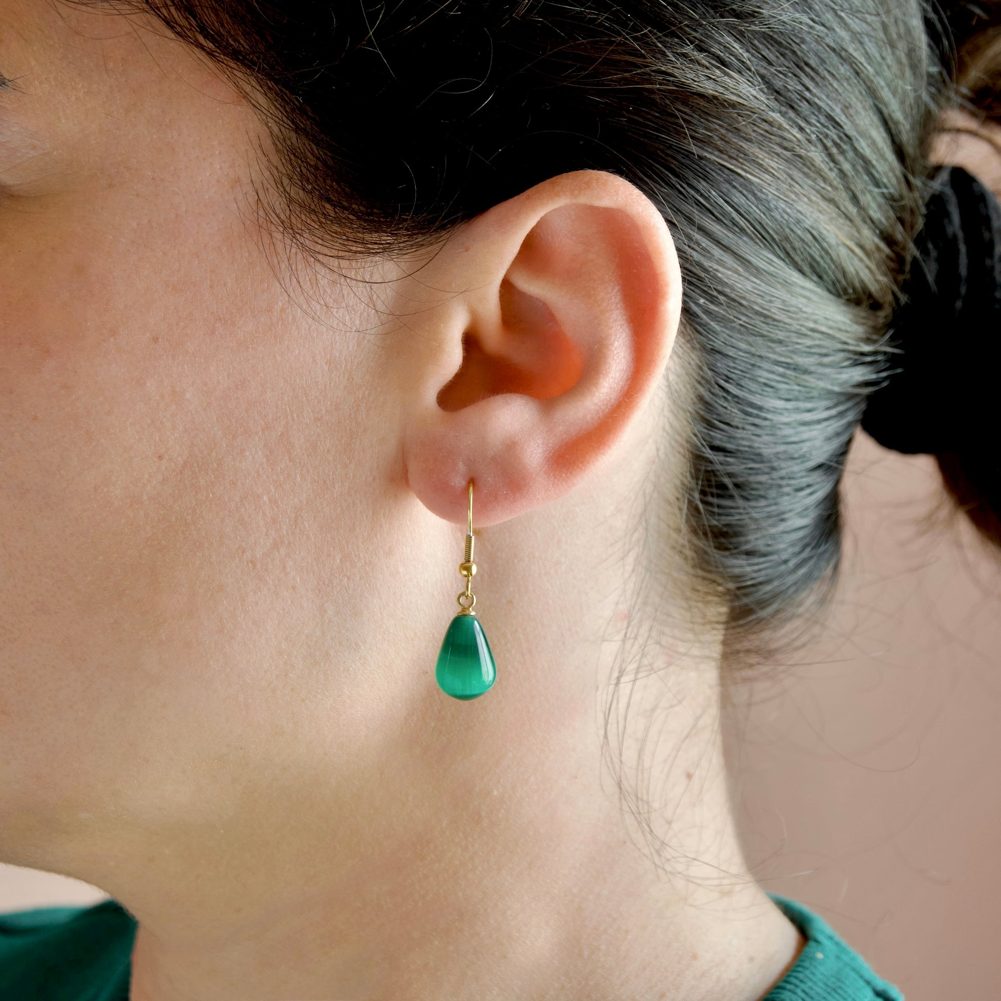 A close-up side profile of a woman wearing a single gold drop earring with a polished green teardrop-shaped stone.