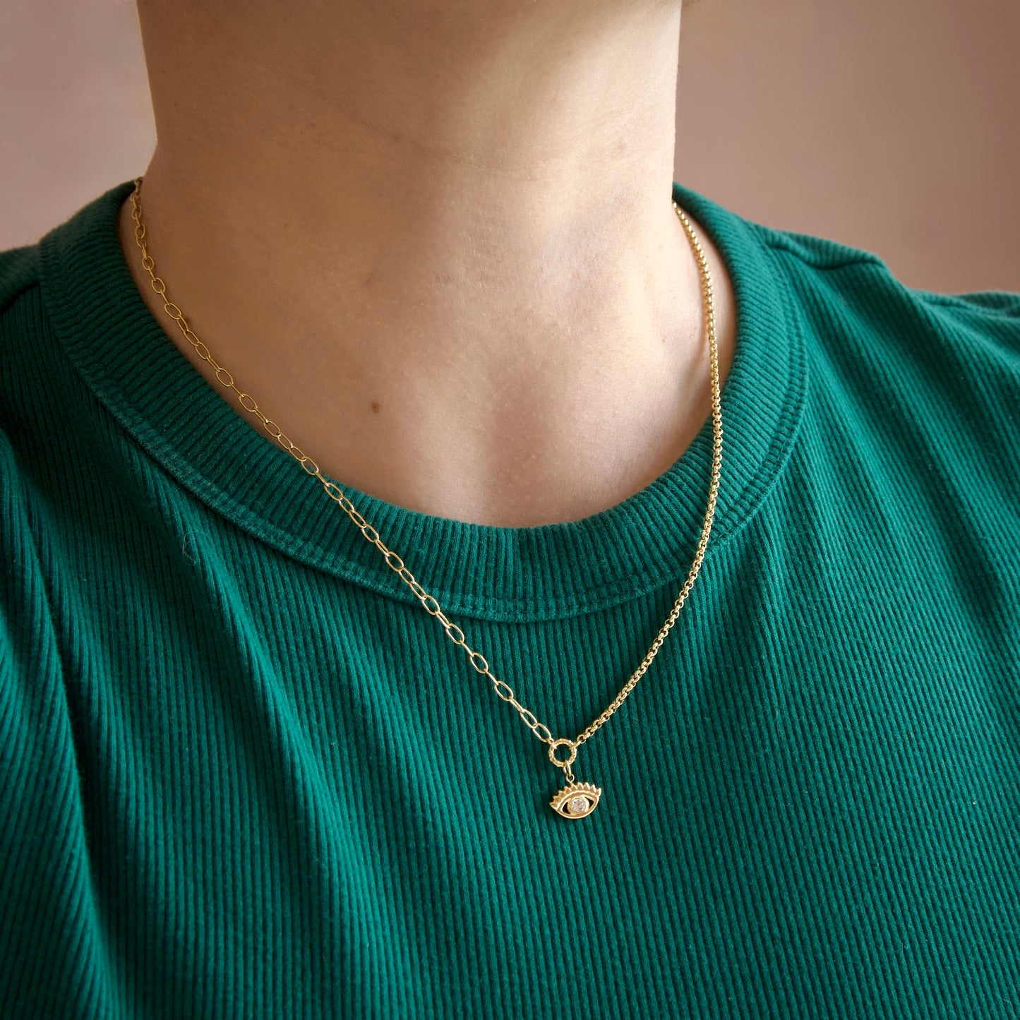 A close-up of a person's neck and upper chest wearing a gold chain necklace with a small detailed pendant, paired with a ribbed green t-shirt against a soft beige background.