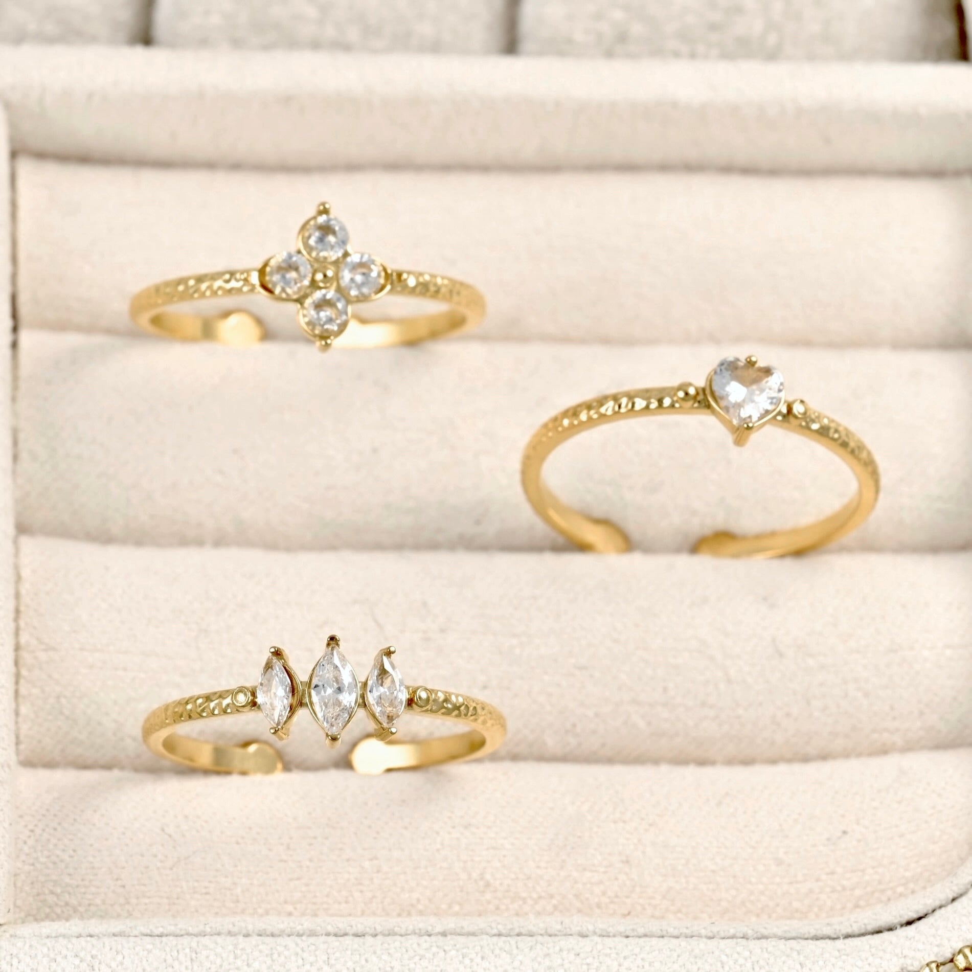 Three gold rings displayed on a beige ring box: one with a four-stone floral cluster, one with a heart-shaped stone, and one with marquise-cut stones.
