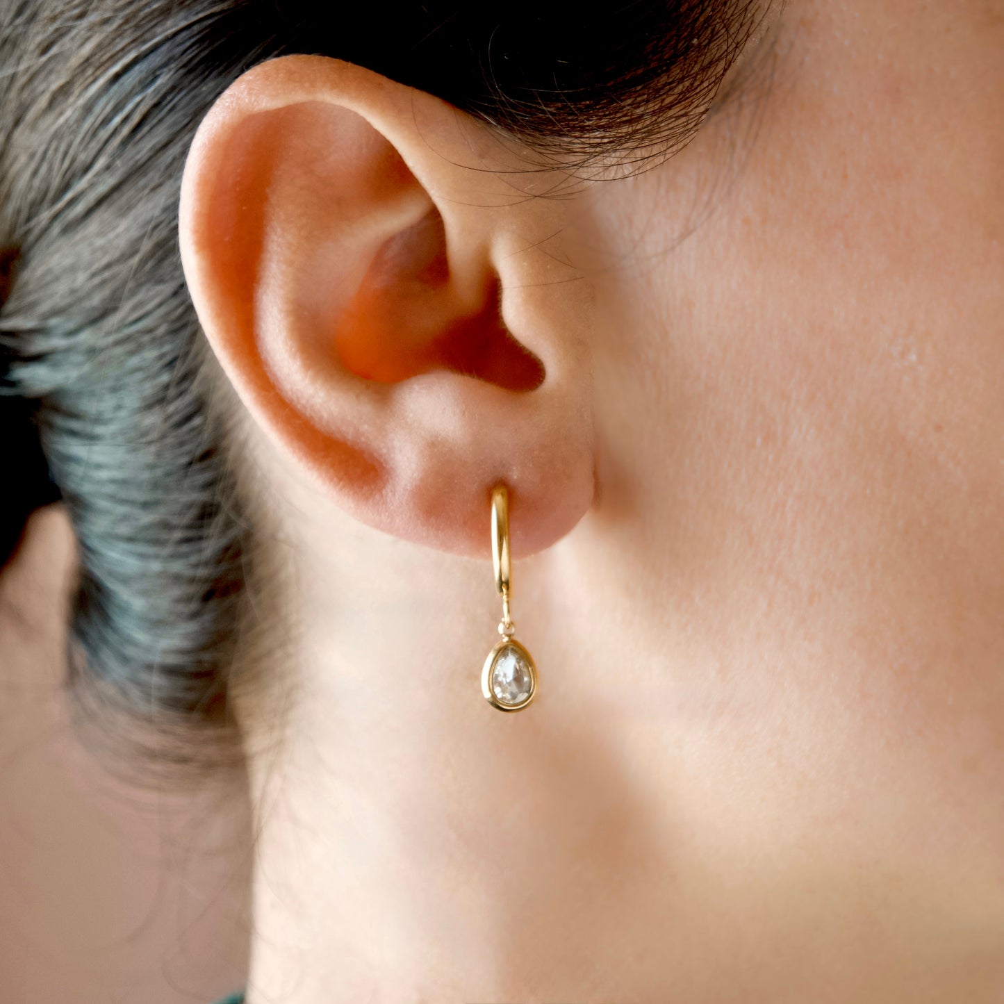 A close-up of a person's ear wearing a delicate gold huggie hoop earring with a small pear cut cubic zirconia dangle.