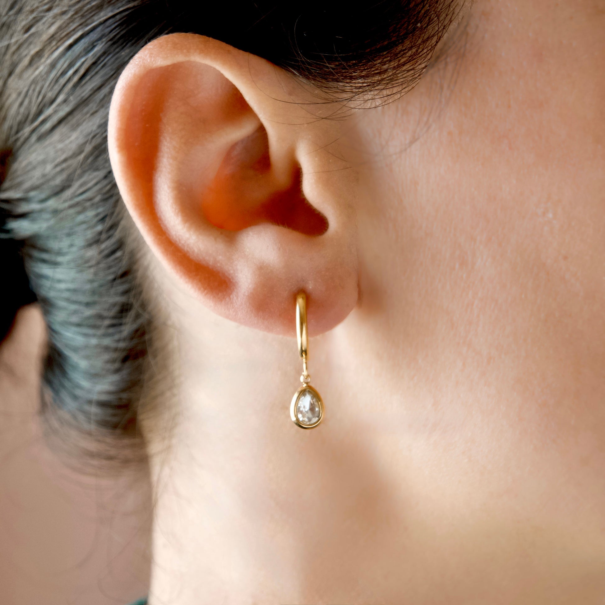A close-up of a person's ear wearing a delicate gold huggie hoop earring with a small pear cut cubic zirconia dangle.