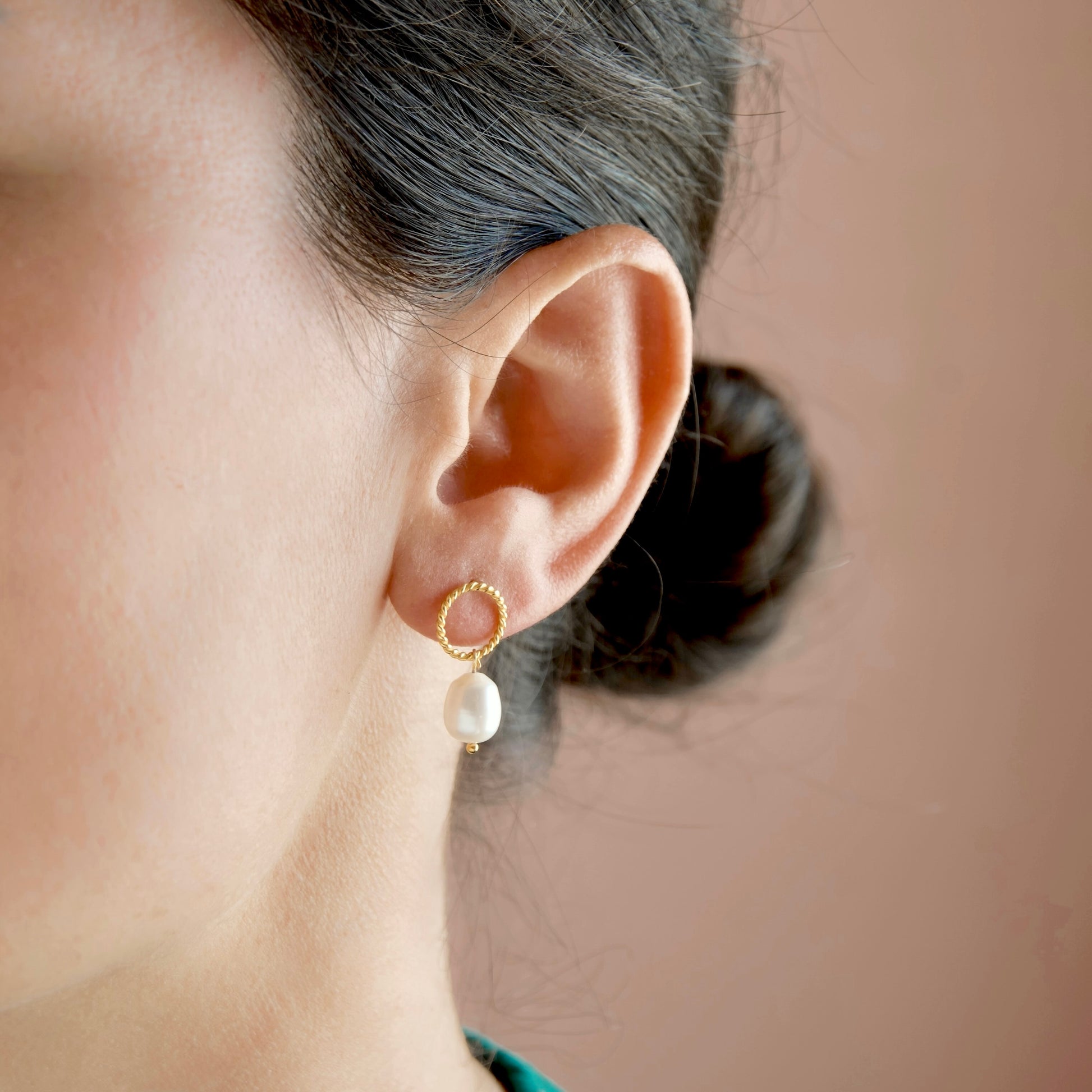 A close-up of a model's ear wearing a gold hoop earring with a dangling white pearl, set against a soft, solid warm brown background.