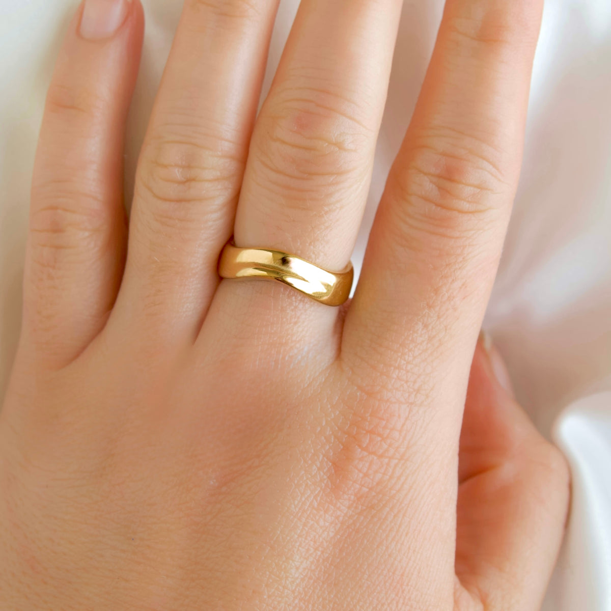 A close-up of a hand wearing a simple polished gold band ring on one of the fingers, set against a soft white background.