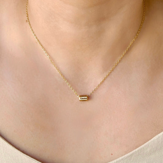A delicate gold chain necklace worn on a person's neck, featuring a small rectangular gold cylinder pendant engraved with the name 'EMMA'.