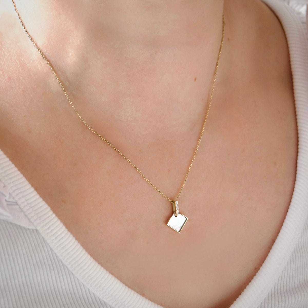 A delicate gold chain necklace with a small, polished mother of pearl square pendant, worn on a model's neck over a white ribbed top.