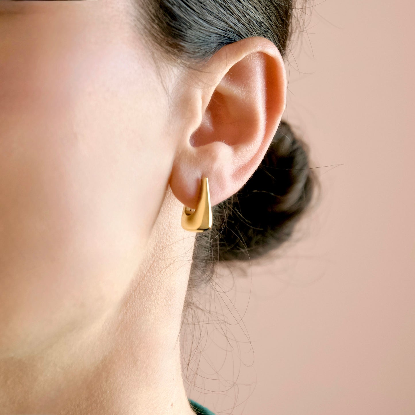 A close-up of a person's ear wearing a small, polished gold geometric stud earring with a triangular top, set against a soft, solid peach-pink background.