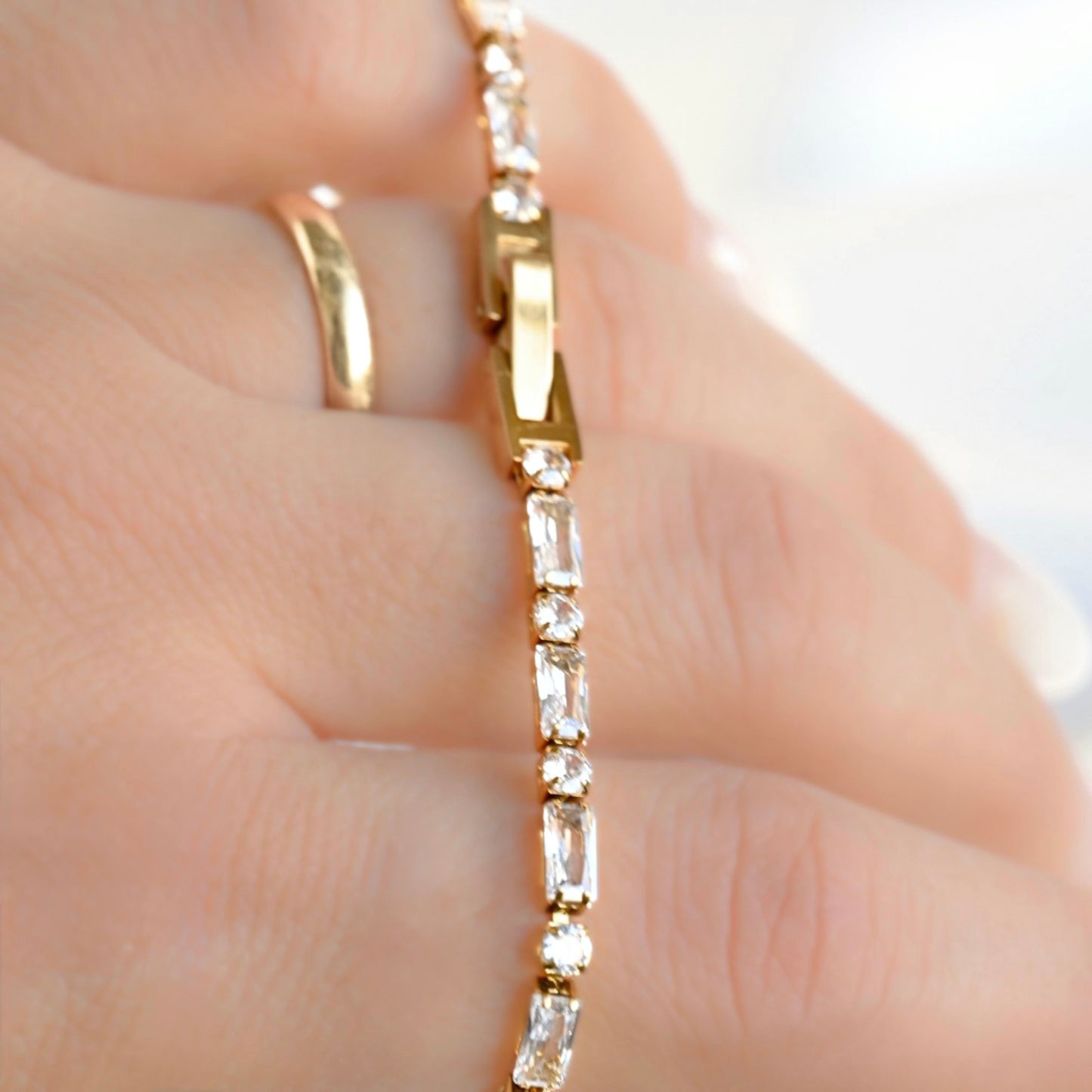 A close-up of a gold bracelet with clear rectangular gemstones worn on a hand, alongside a simple gold ring.