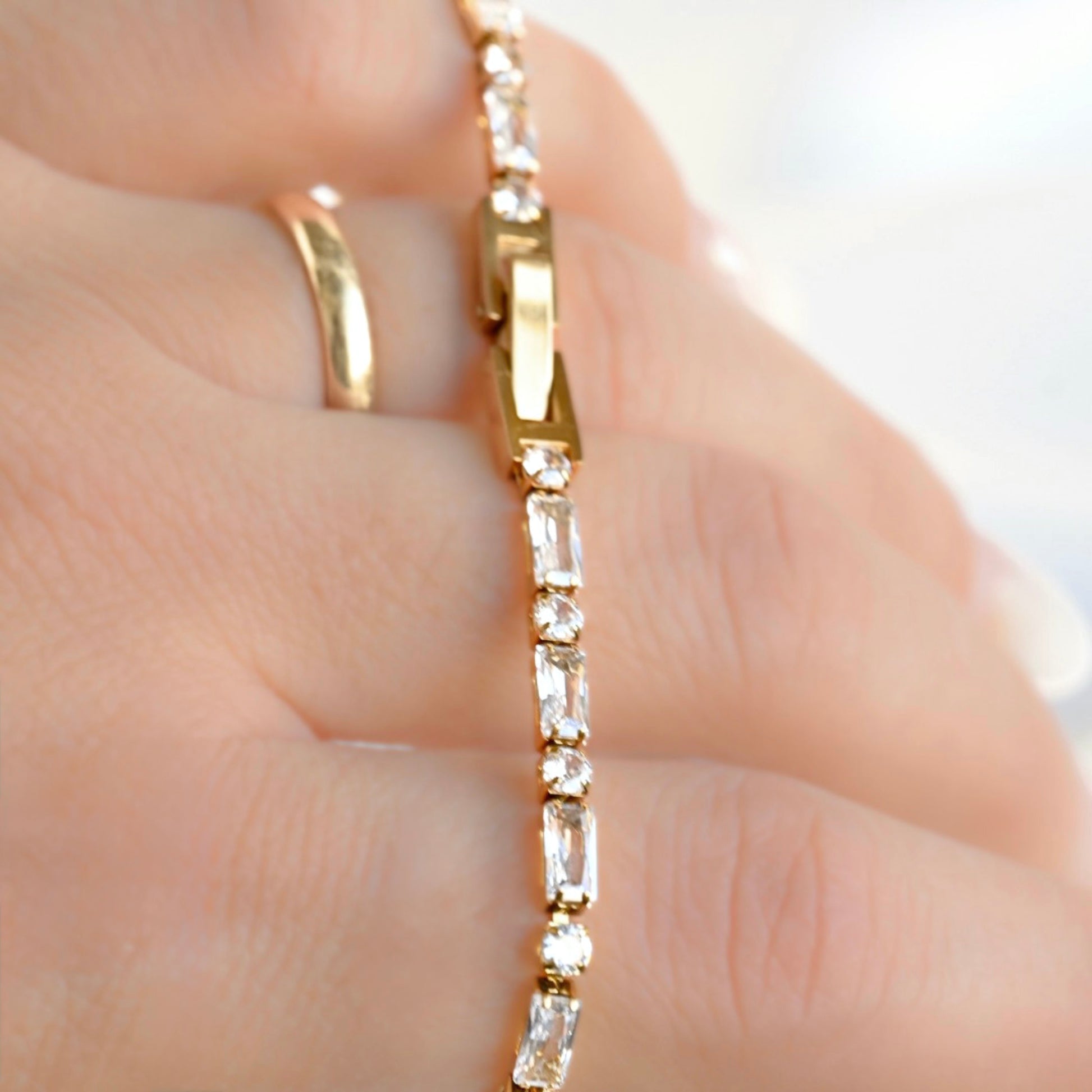 A close-up of a gold bracelet with clear rectangular gemstones worn on a hand, alongside a simple gold ring.