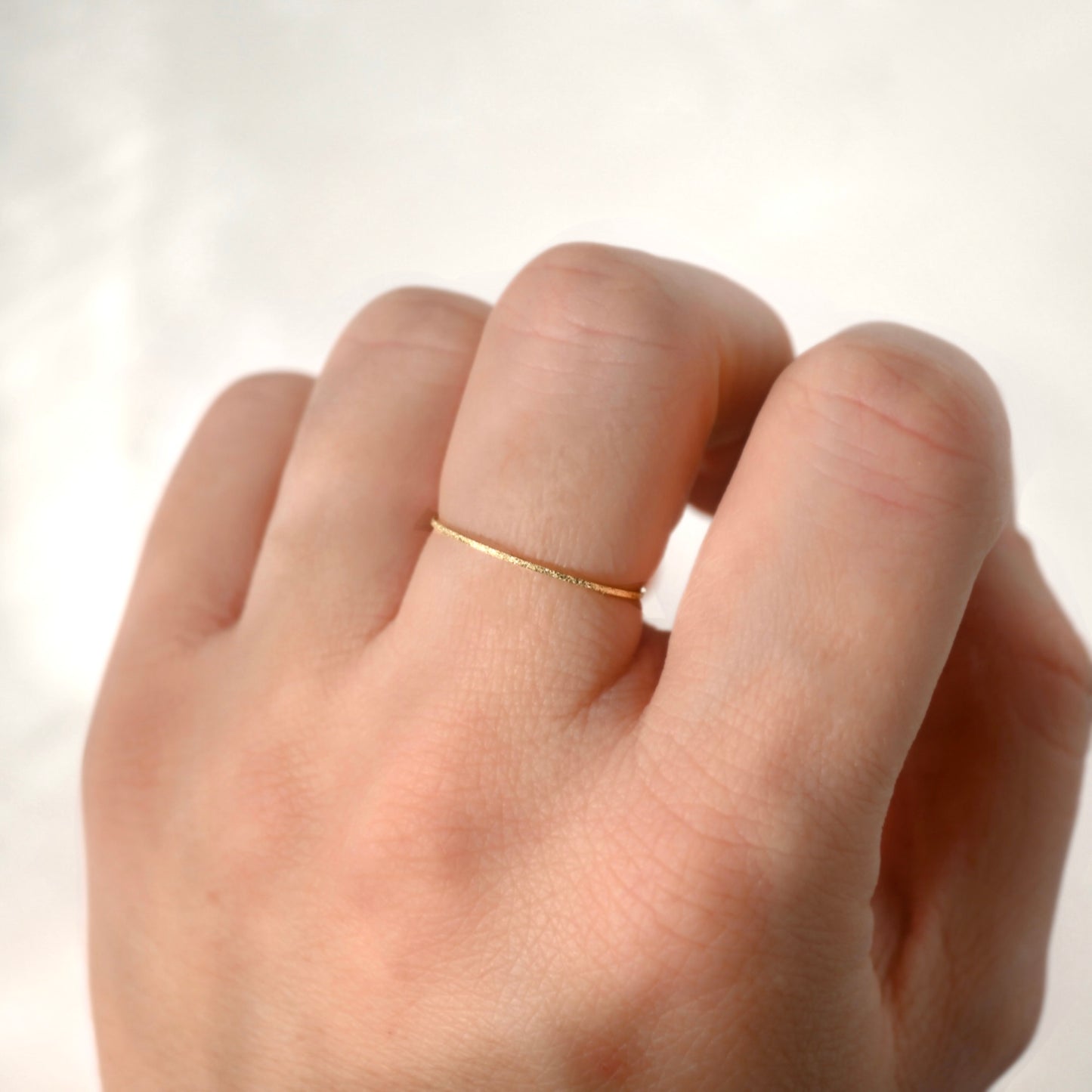 A close-up of a hand wearing a thin, delicate gold band ring on one of the fingers, set against a soft, out-of-focus light background.
