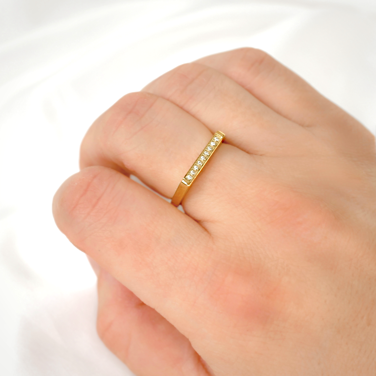 A close-up of a hand wearing a slim yellow gold ring with a row of small clear stones on the ring finger, set against a soft white background.