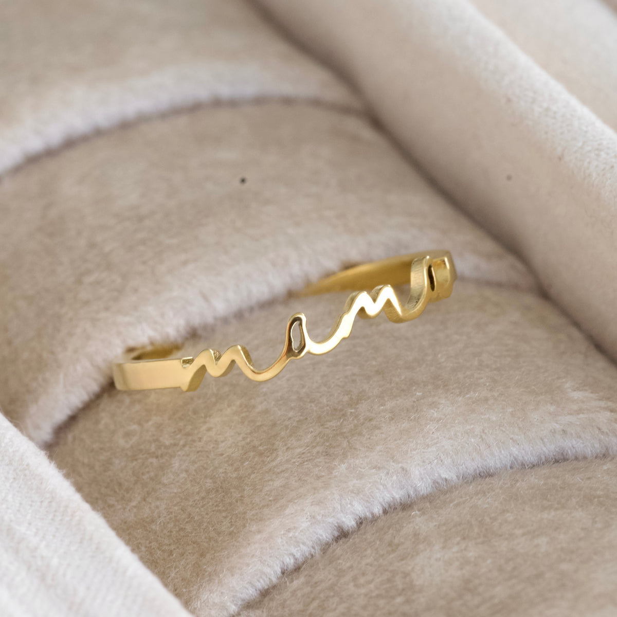 A gold ring with a wavy, flowing line design with 'mama' engraving, displayed on a beige textured velvet ring box.
