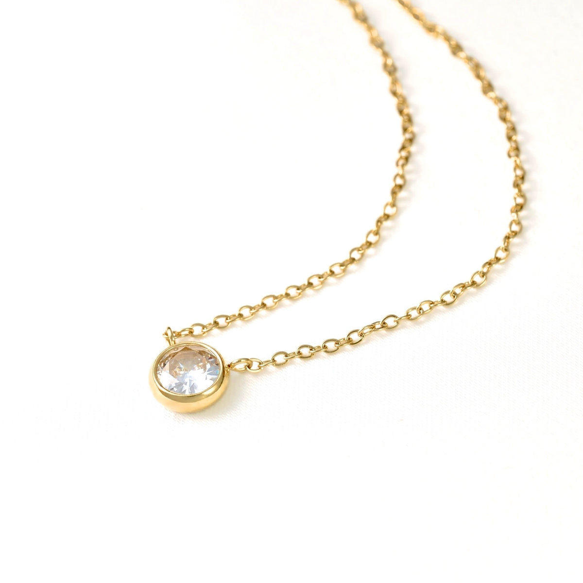 A delicate gold chain necklace featuring a small round bezel-set clear gemstone pendant, photographed on a clean white background.