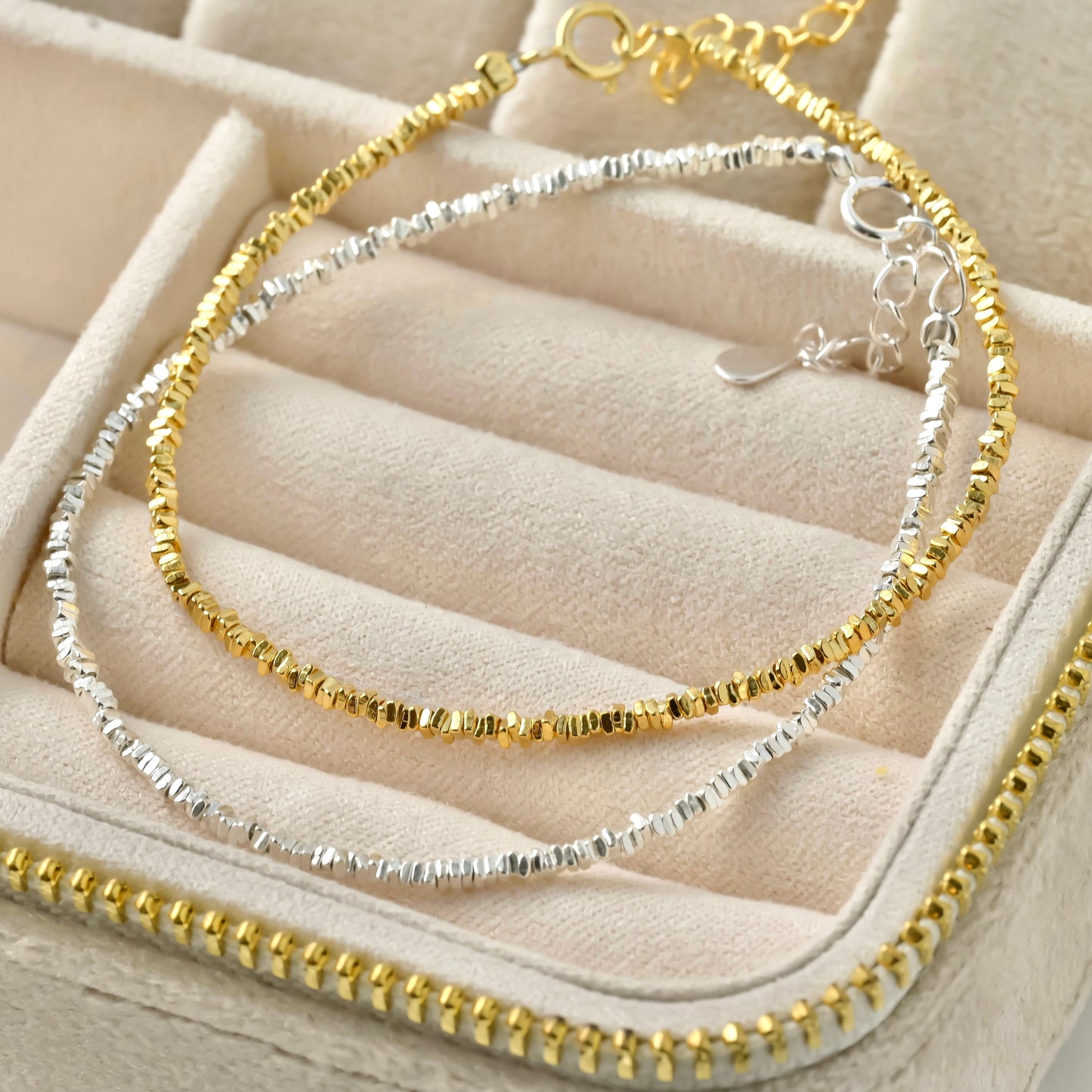 Two gold and silver beaded bracelets on a beige jewelry display.