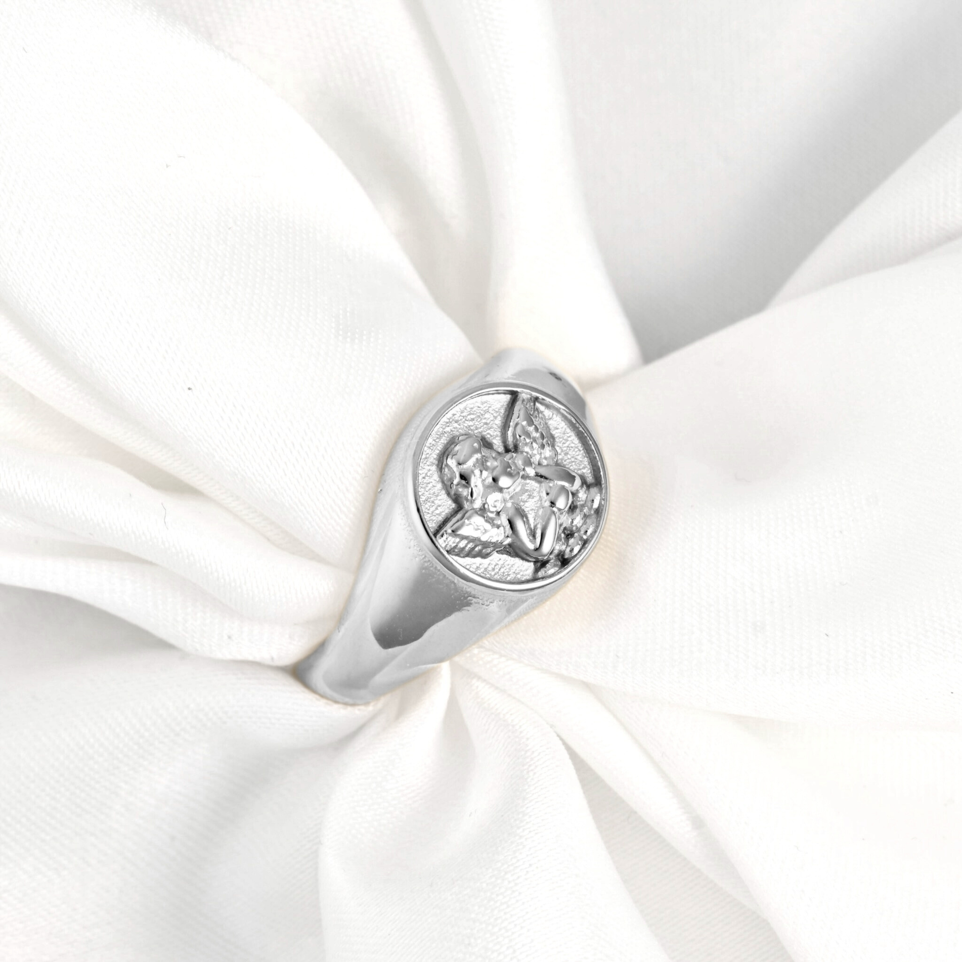 A silver signet ring featuring an engraved angel design, displayed on a soft, white draped fabric background.